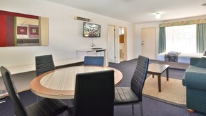 A hotel room with a round dining table, black chairs, a sofa, a TV on the wall, and a bed with a window view. - Coffs Windmill Motel (Coffs Harbour)