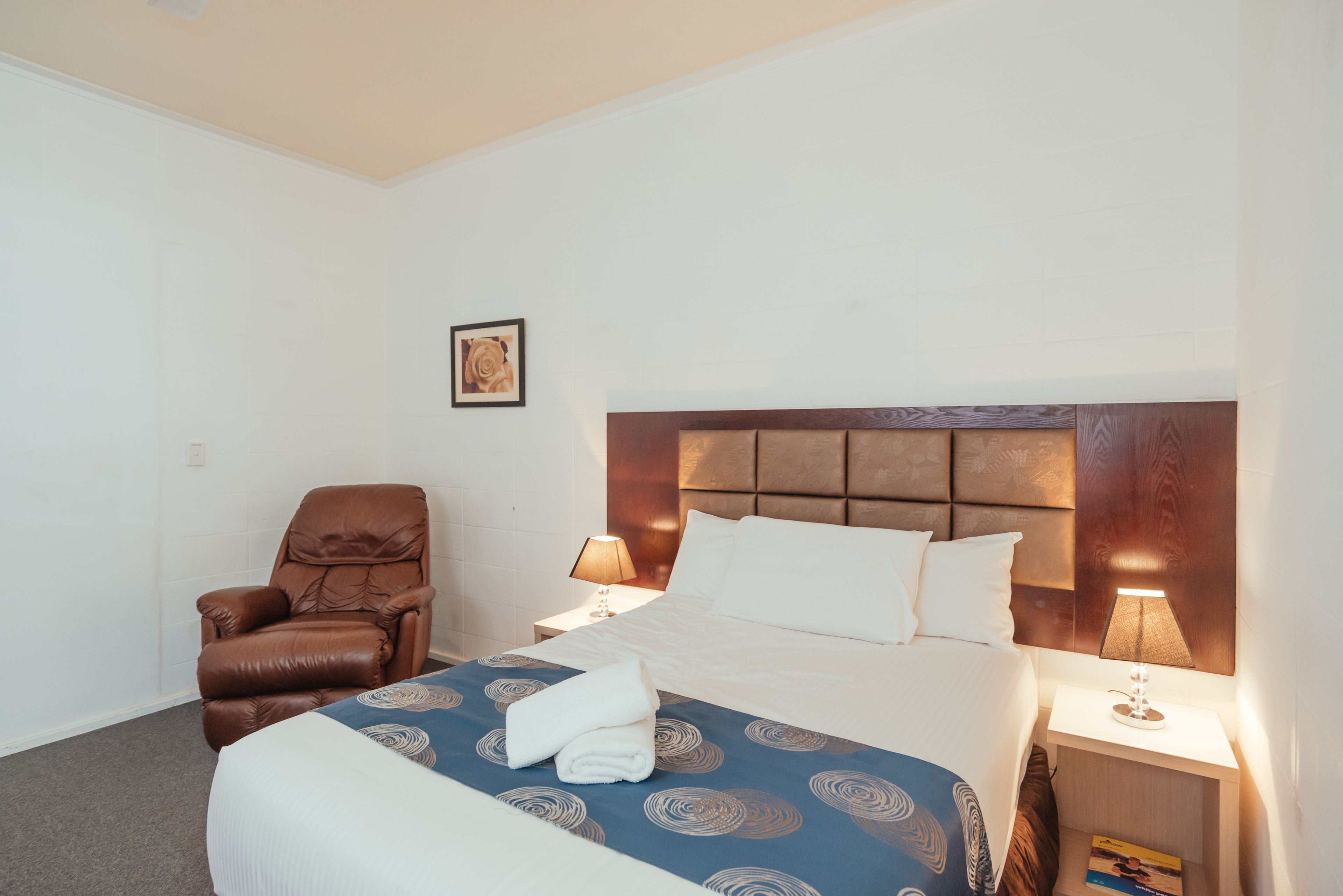 A hotel room with a bed, a brown leather chair, a nightstand with a lamp, and a framed picture on the wall.