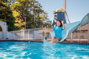A swimming pool with a water slide, surrounded by a fence and trees. - Coffs Windmill Motel (Coffs Harbour)