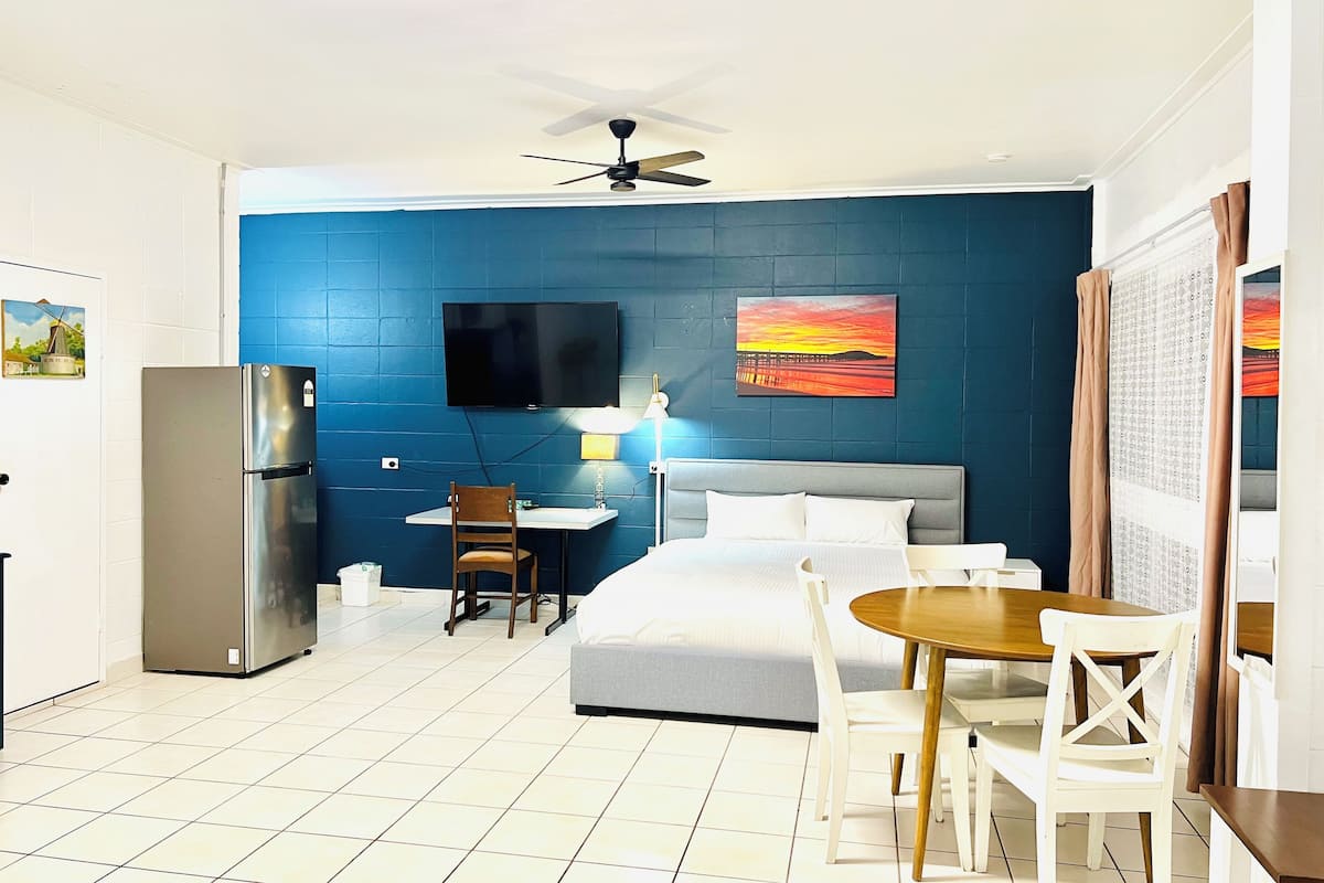 A hotel room with a bed, a desk with a chair, a refrigerator, a ceiling fan, and a TV mounted on the wall.