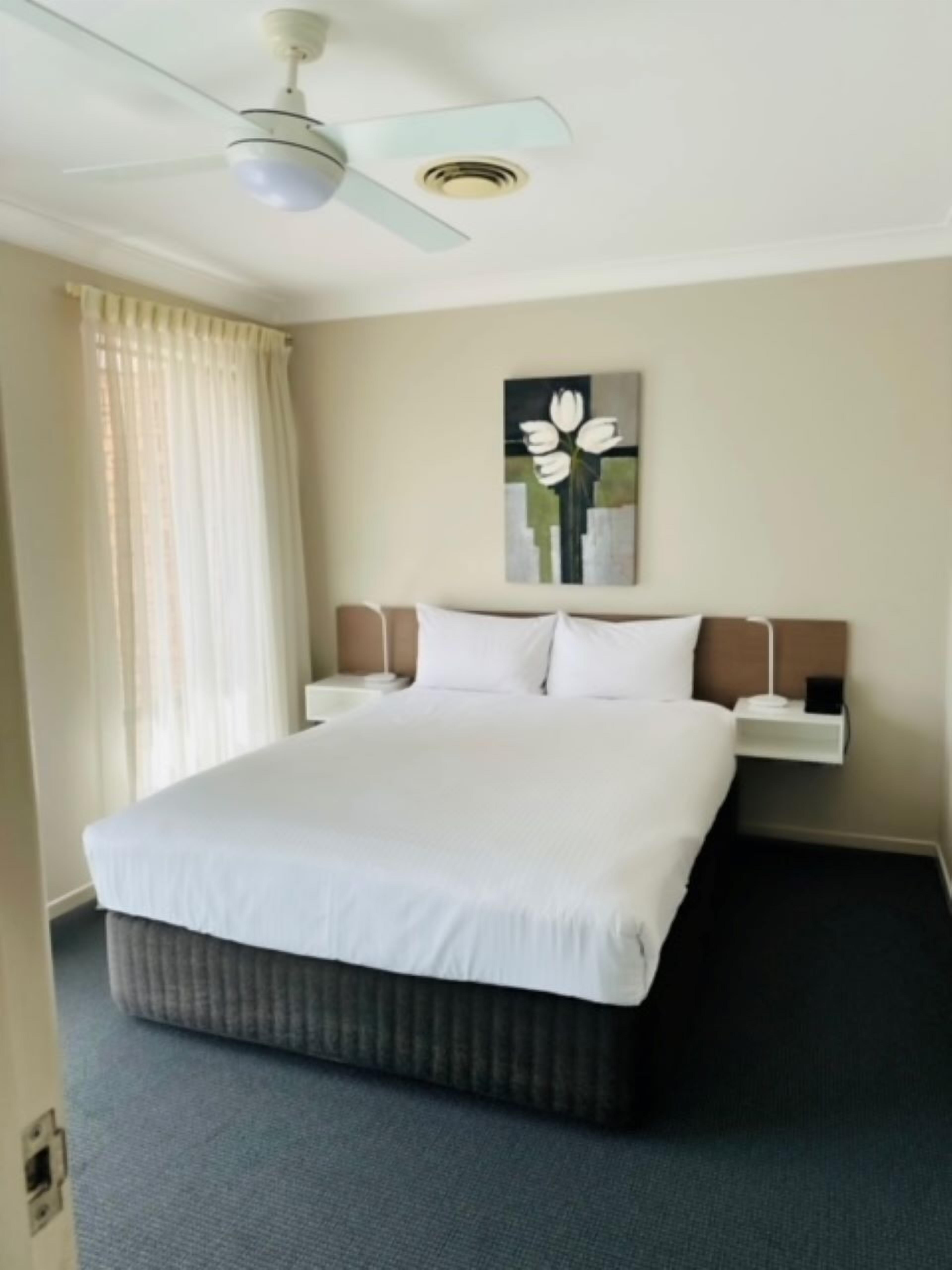 Une chambre d’hôtel avec un grand lit, un ventilateur de plafond, un tableau représentant des fleurs et deux tables de chevet avec des lampes.
