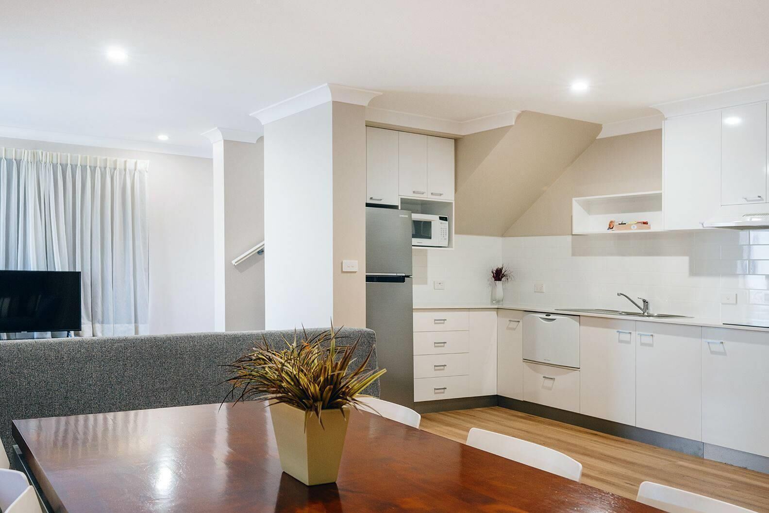 A modern kitchen with white cabinets, a microwave, and a sink. There is a dining table with a plant in the center.