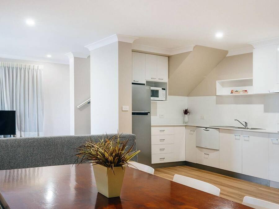 A modern kitchen with white cabinets, a microwave, and a sink. There is a dining table with a plant in the center.