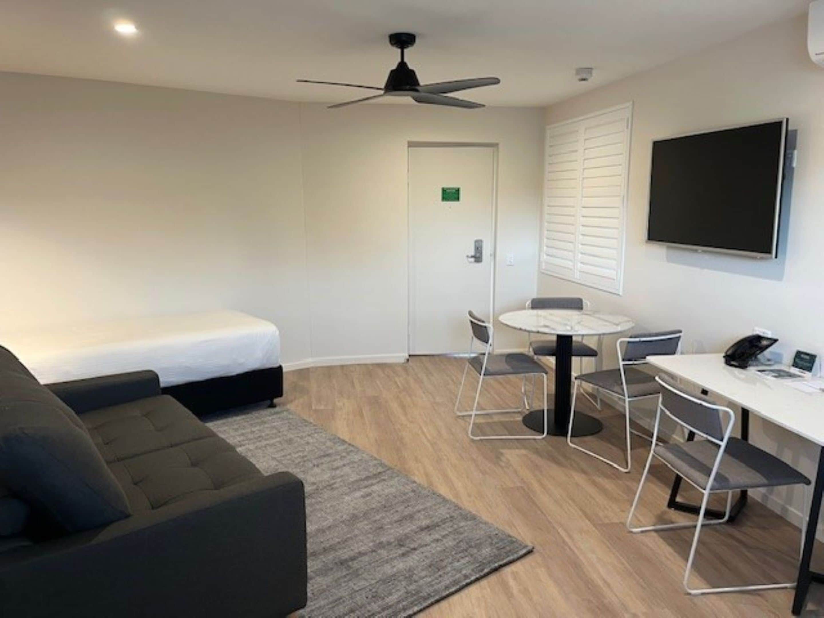 A modern hotel room with a bed, a sofa, a desk with chairs, a ceiling fan, and a TV.
