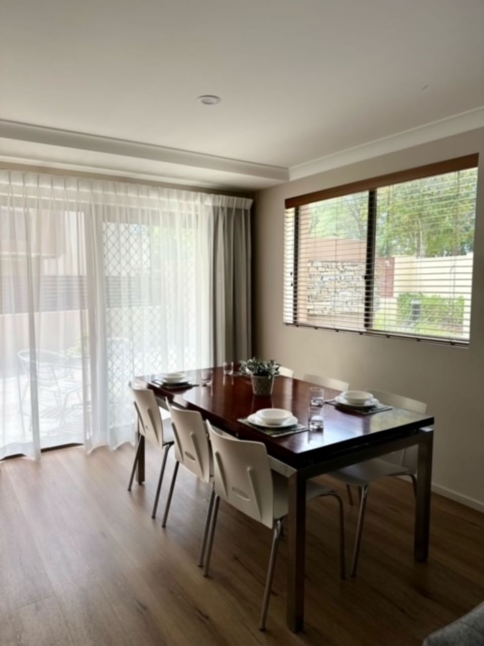 A dining area with a wooden table, white chairs, and a window with blinds.