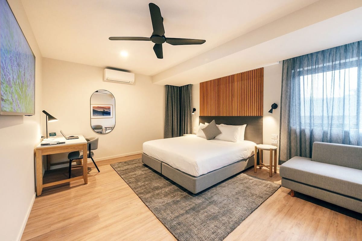 A hotel room with a bed, a desk, a chair, a sofa, a ceiling fan, and a window with curtains.