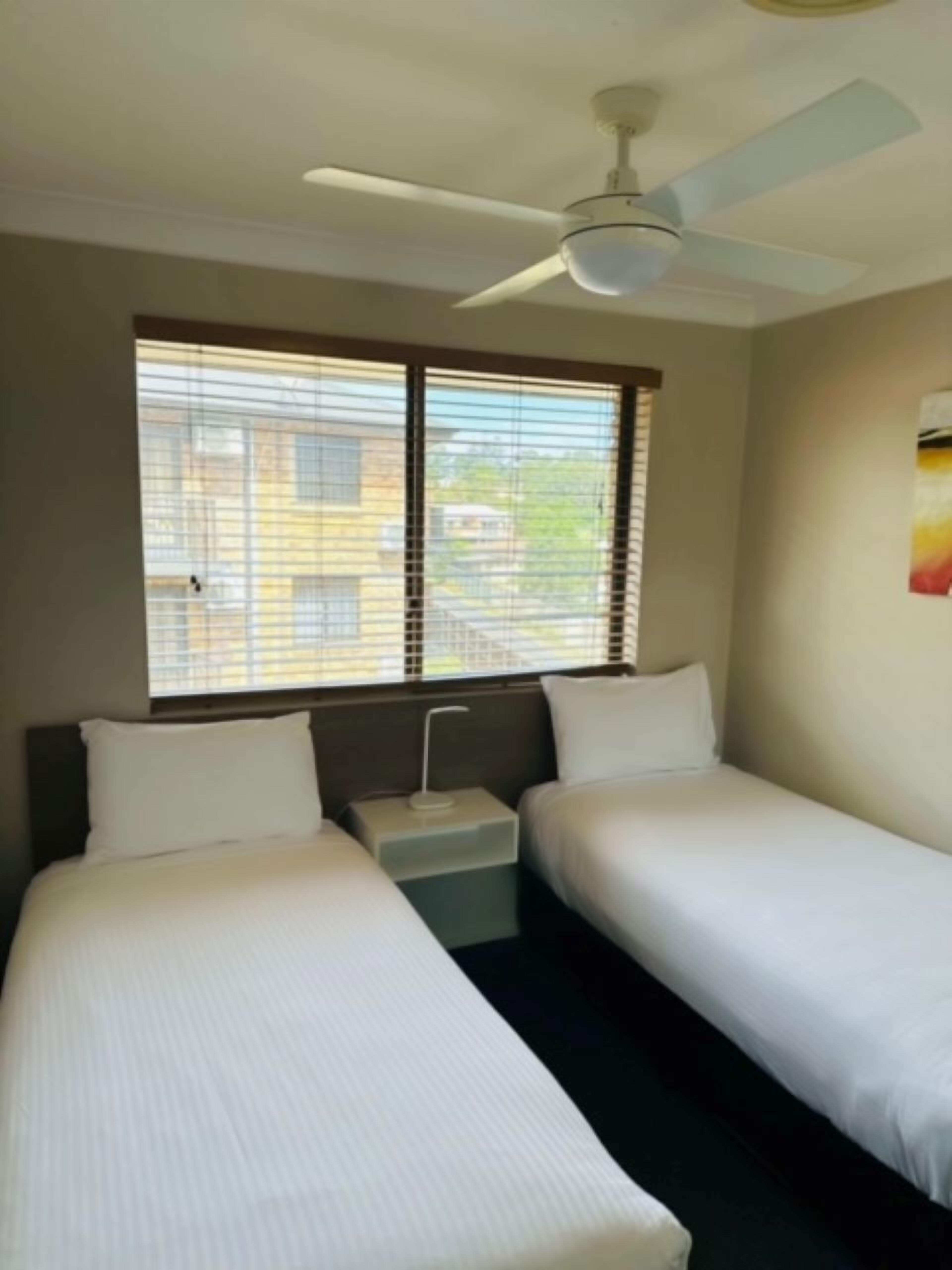 Quarto de hotel com duas camas de solteiro, um ventilador de teto e janela com persianas.