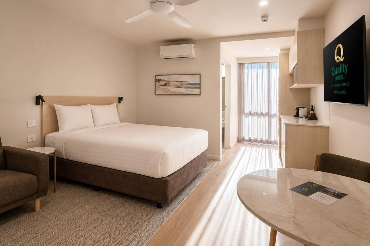 Habitación de hotel con cama, escritorio, silla, ventilador de techo y televisor mostrando el nombre del hotel.