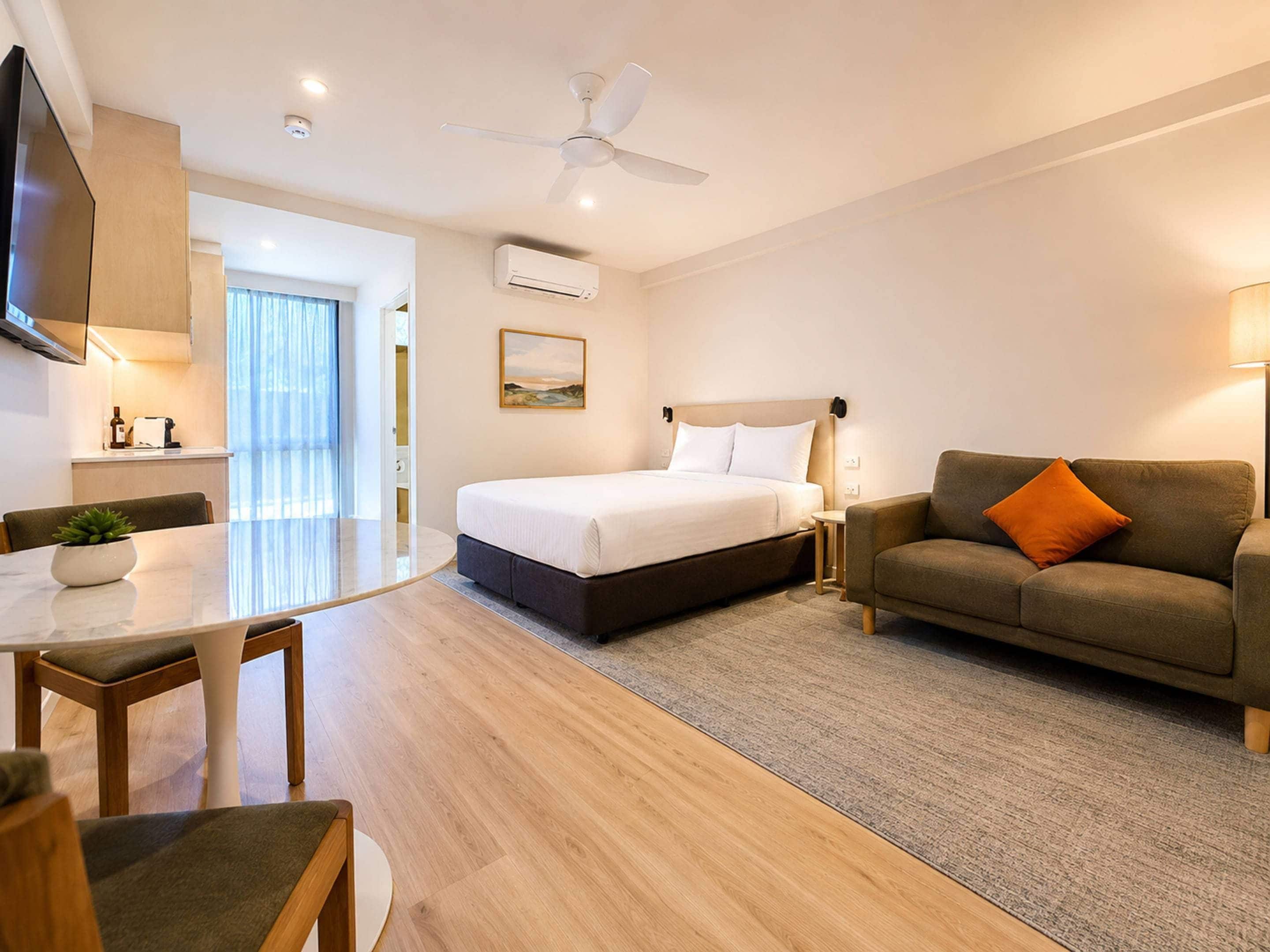 A modern hotel room with a bed, a sofa, a dining area, and a ceiling fan.