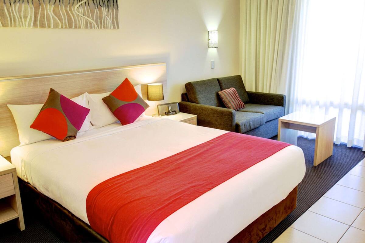 A hotel room with a large bed, a sofa, a nightstand, a lamp, and a view of the outdoors.