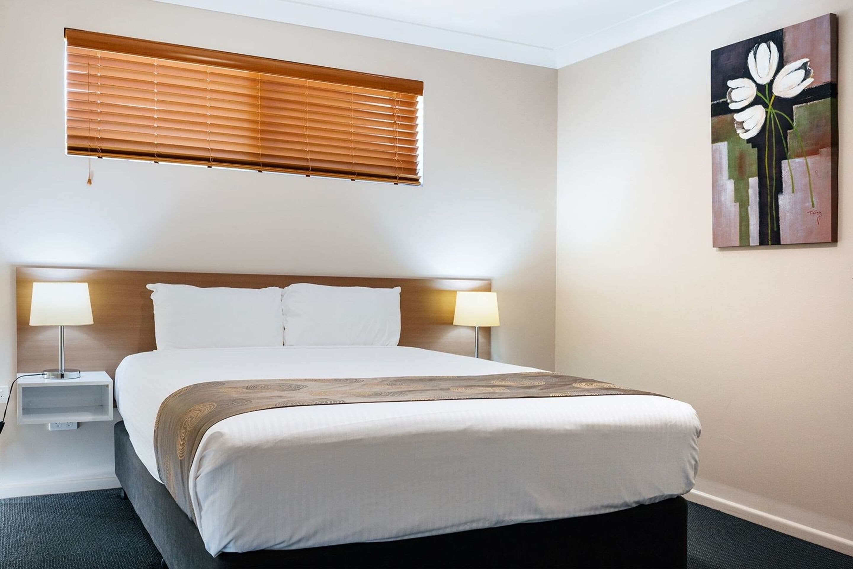 Habitación de hotel con una cama grande, dos mesitas de noche con lámparas, una ventana con persianas y un cuadro en la pared.