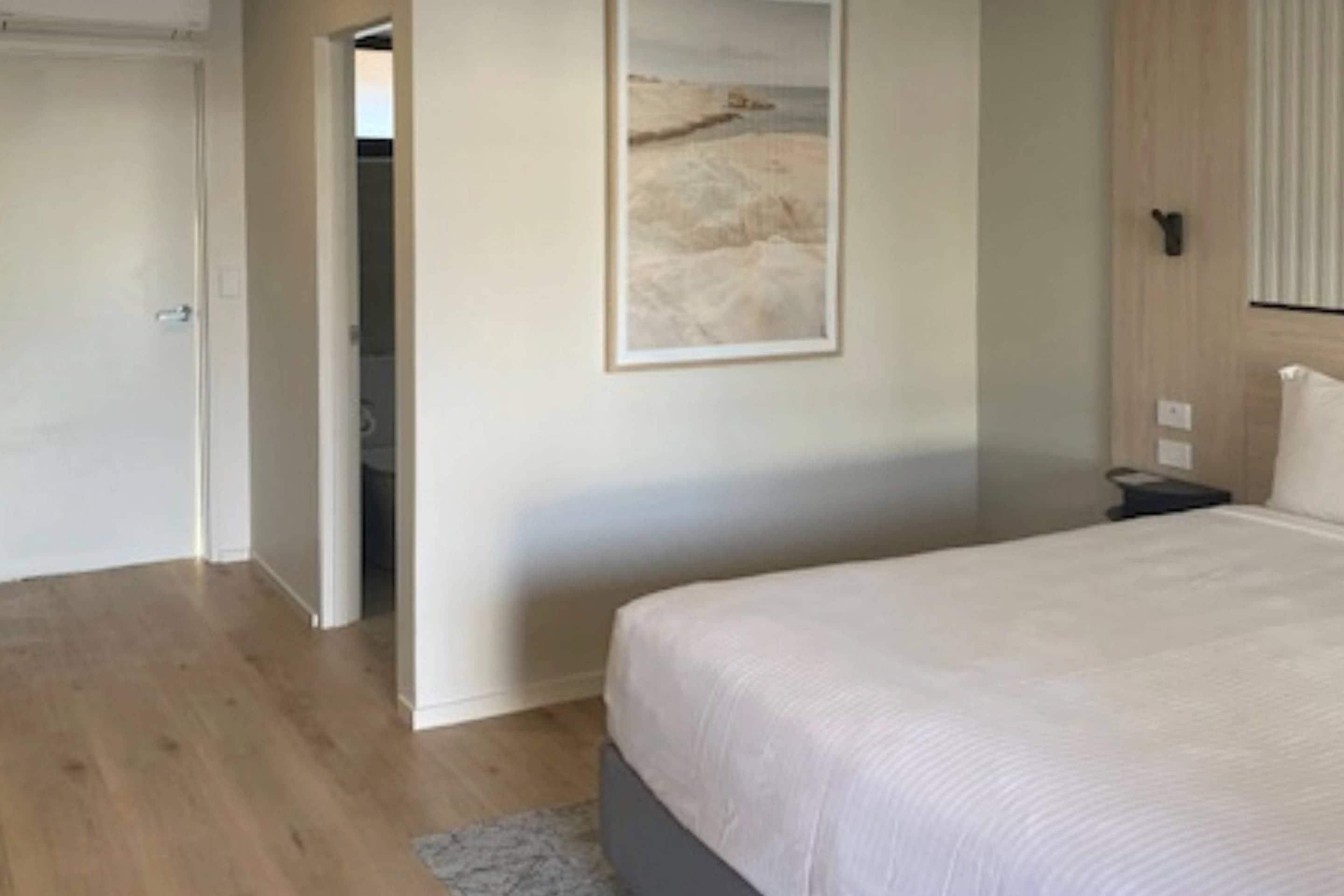 A bedroom with a bed, a framed picture on the wall, and a door leading to another room.
