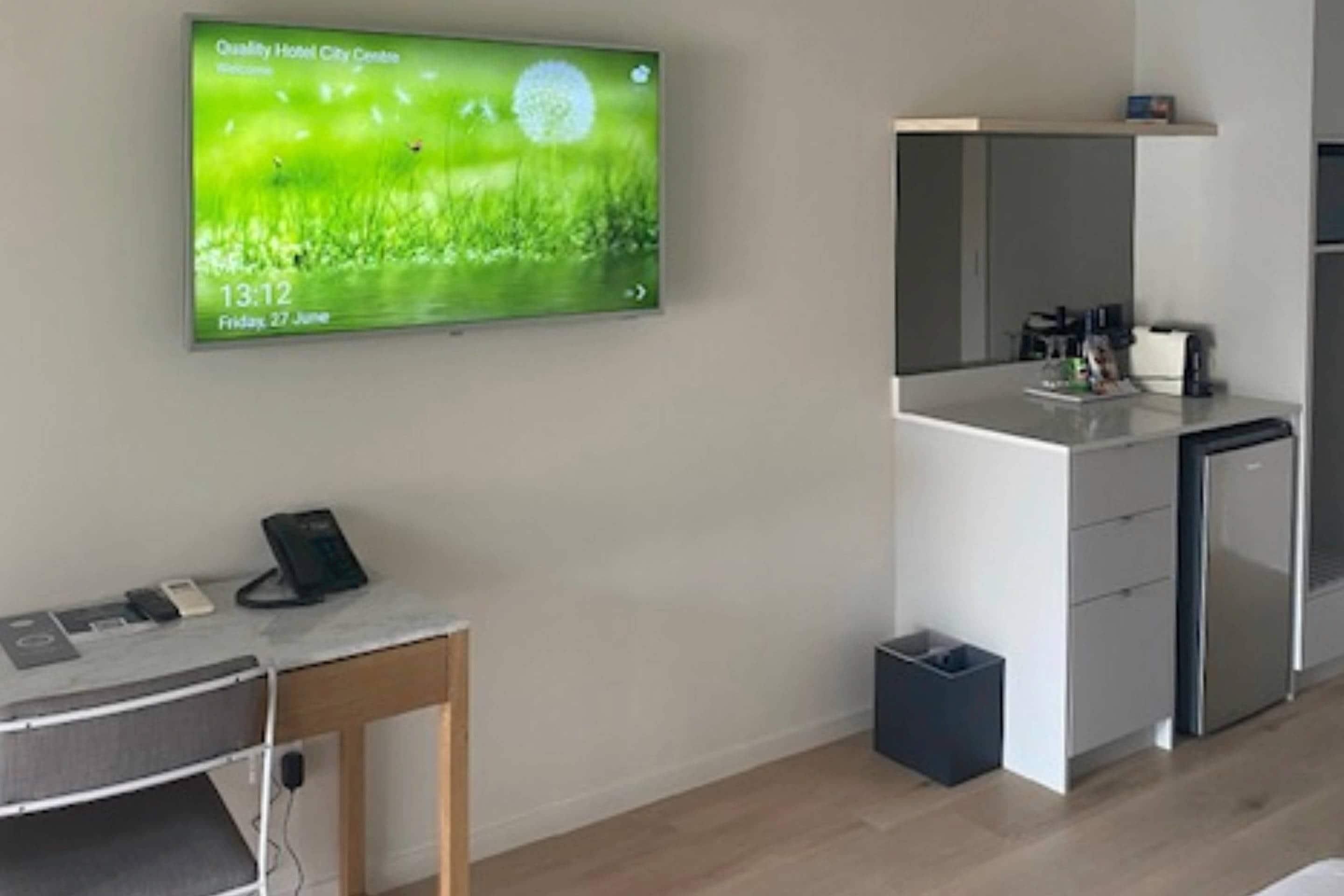 A modern hotel room with a flat-screen TV displaying a nature scene, a desk with a phone, a mirror, and a small kitchenette.