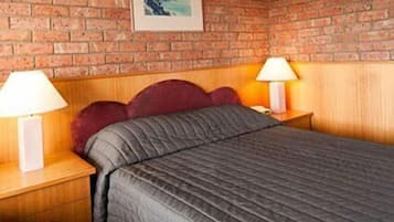 A room with a brick wall, a bed with a dark quilt, two bedside tables with lamps, and a framed picture on the wall.