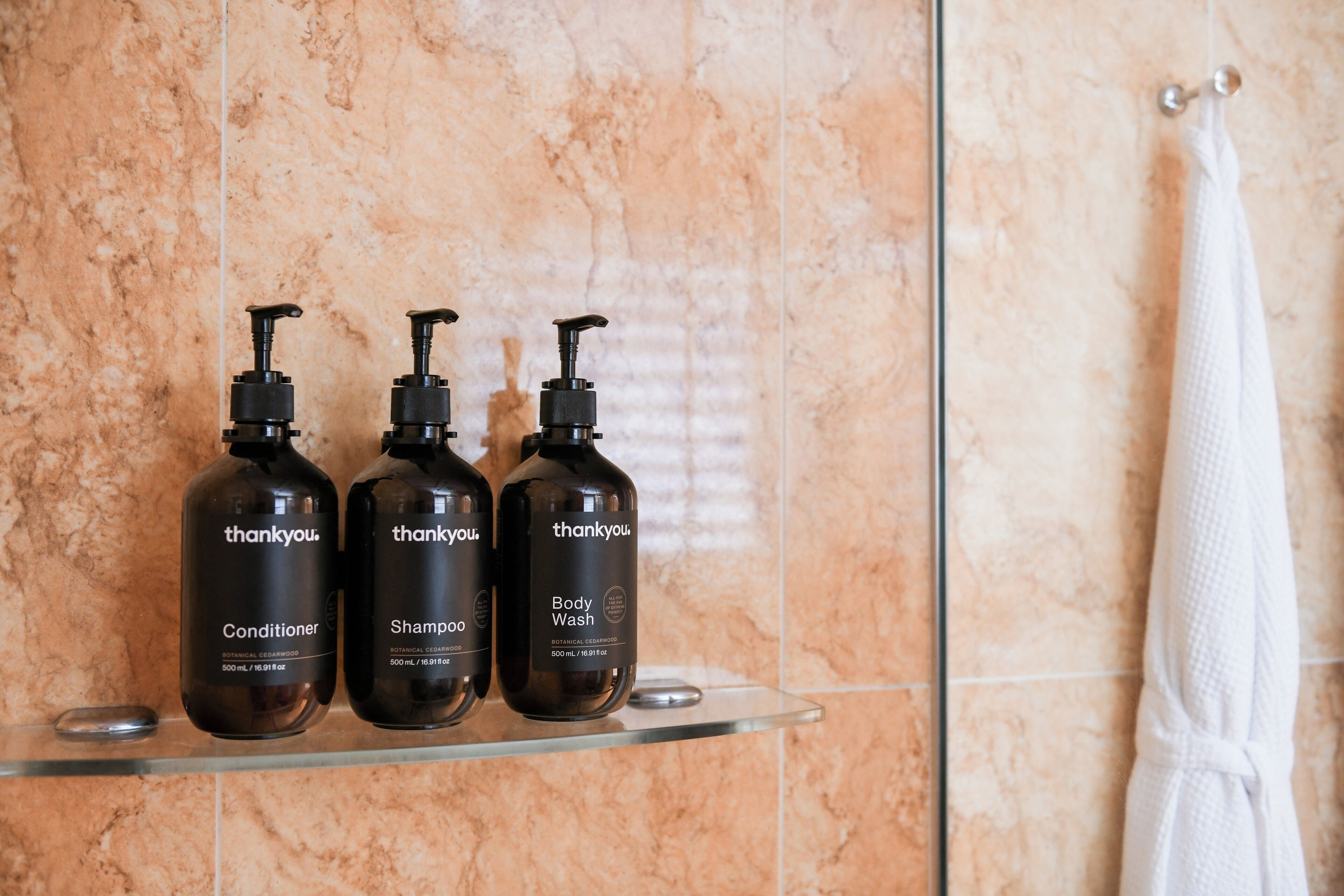 Trois bouteilles de produits de soins corporels de la marque « thankyou » sont posées sur une étagère en verre dans une salle de bain dont le mur est en marbre et où une serviette blanche est suspendue à un porte-serviettes.
