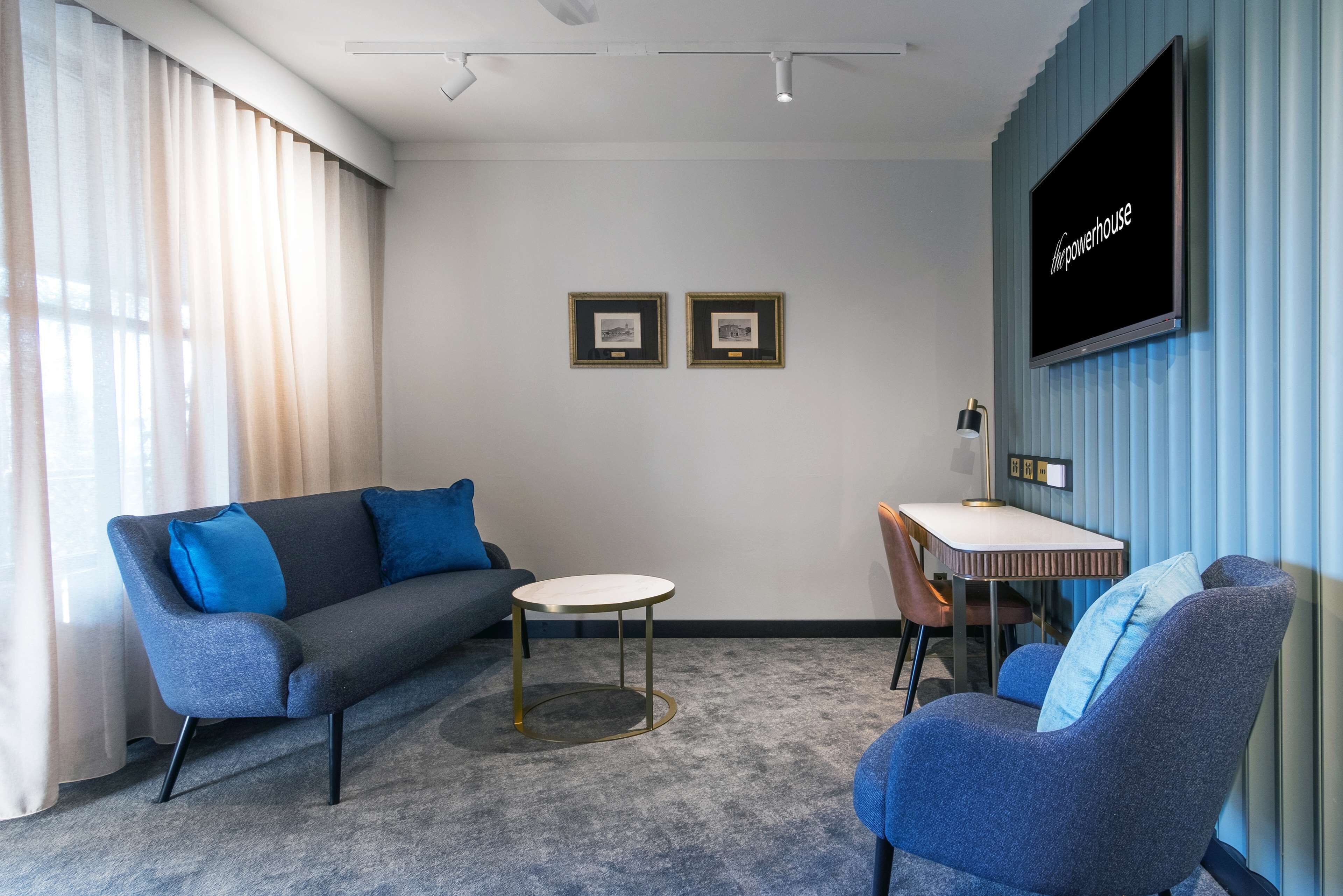 A modern hotel room with a grey sofa, a blue armchair, a small round table, and a TV mounted on a blue wall.
