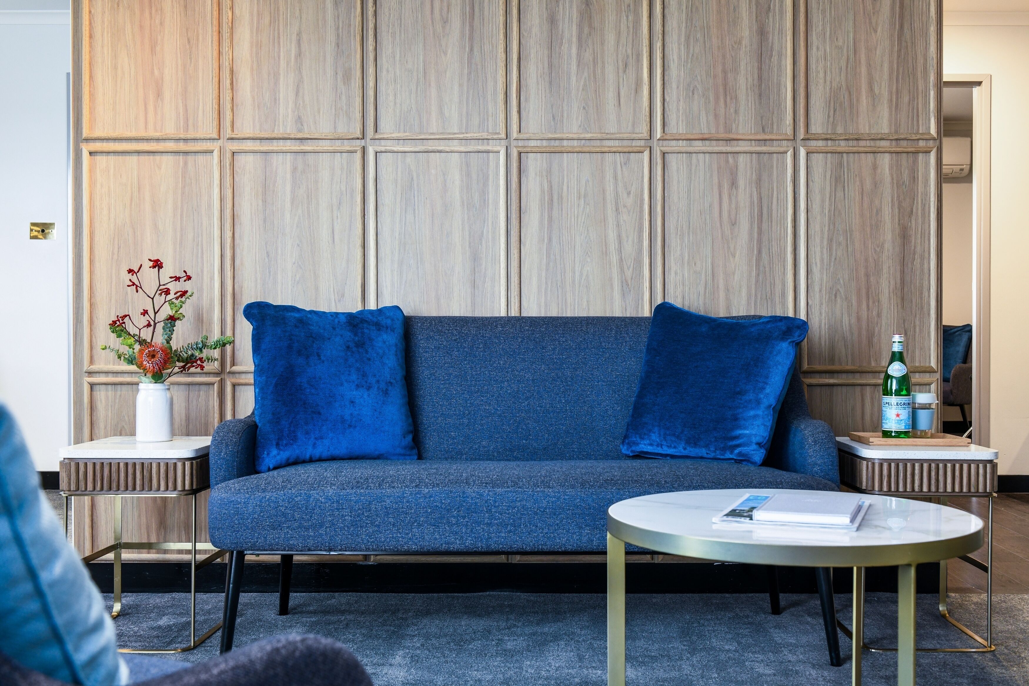 Un salon moderne avec un canapé bleu, deux coussins assortis, une petite table d’appoint blanche, une table basse ronde et un vase décoratif avec des fleurs.