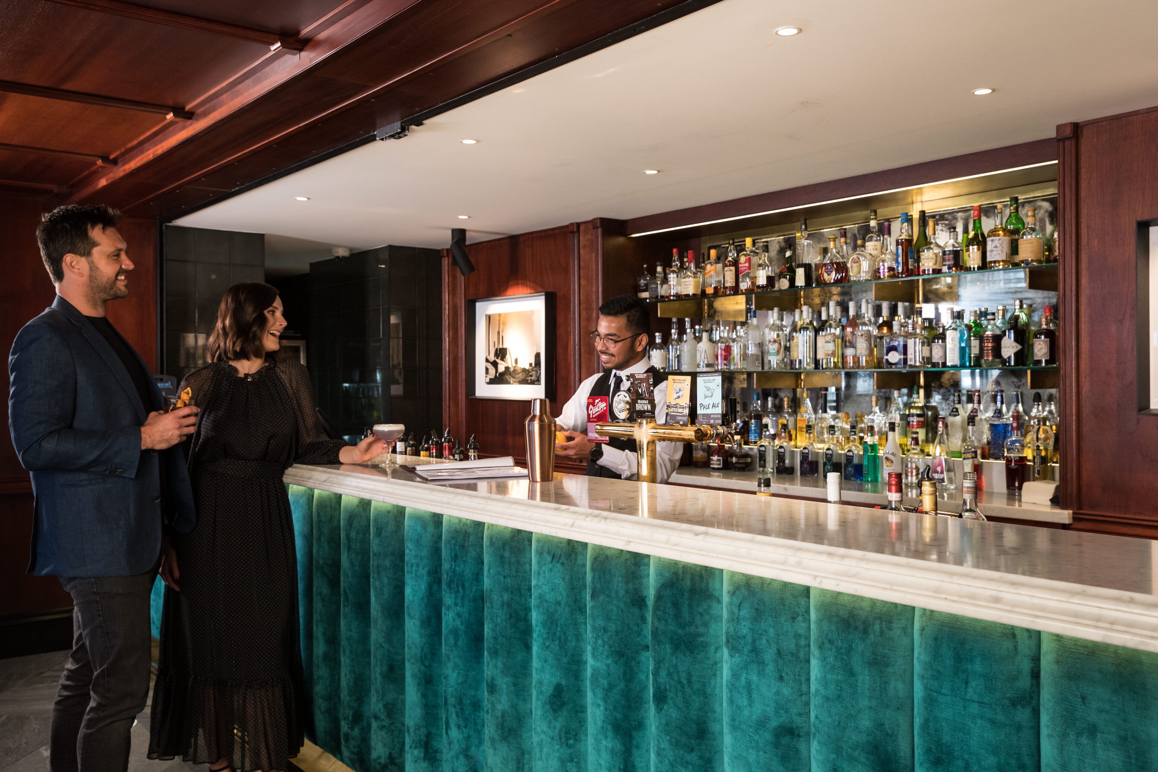 A bar area with a bartender, a counter with a teal velvet backrest, and shelves stocked with various liquor bottles.