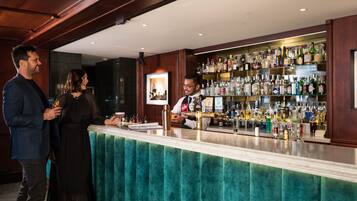 A bar area with a bartender, a counter with a teal velvet backrest, and shelves stocked with various liquor bottles.