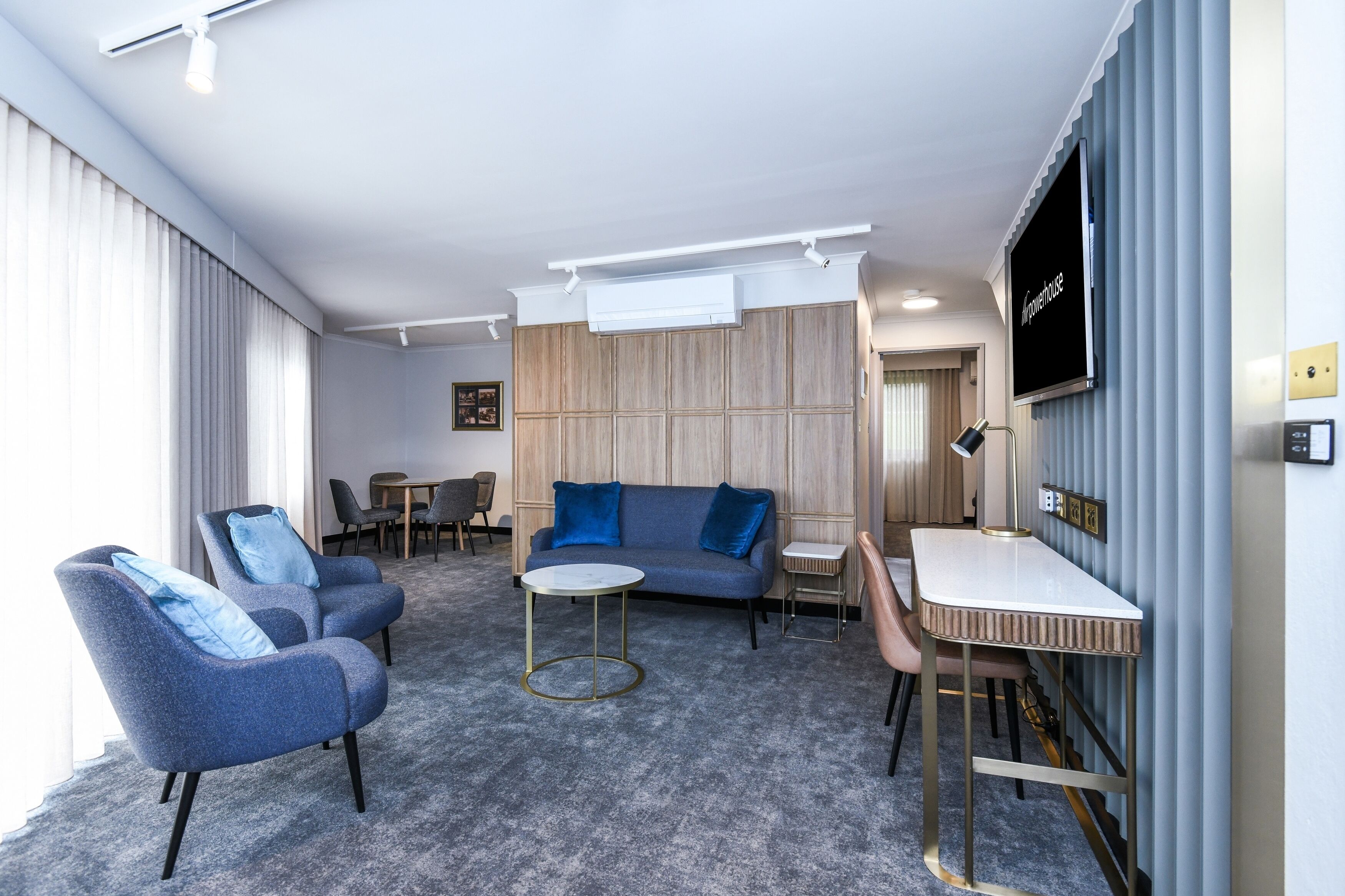Une chambre d’hôtel moderne avec un tapis gris, un canapé bleu, deux fauteuils, une petite table ronde, un téléviseur fixé au mur et un mur recouvert de panneaux en bois.