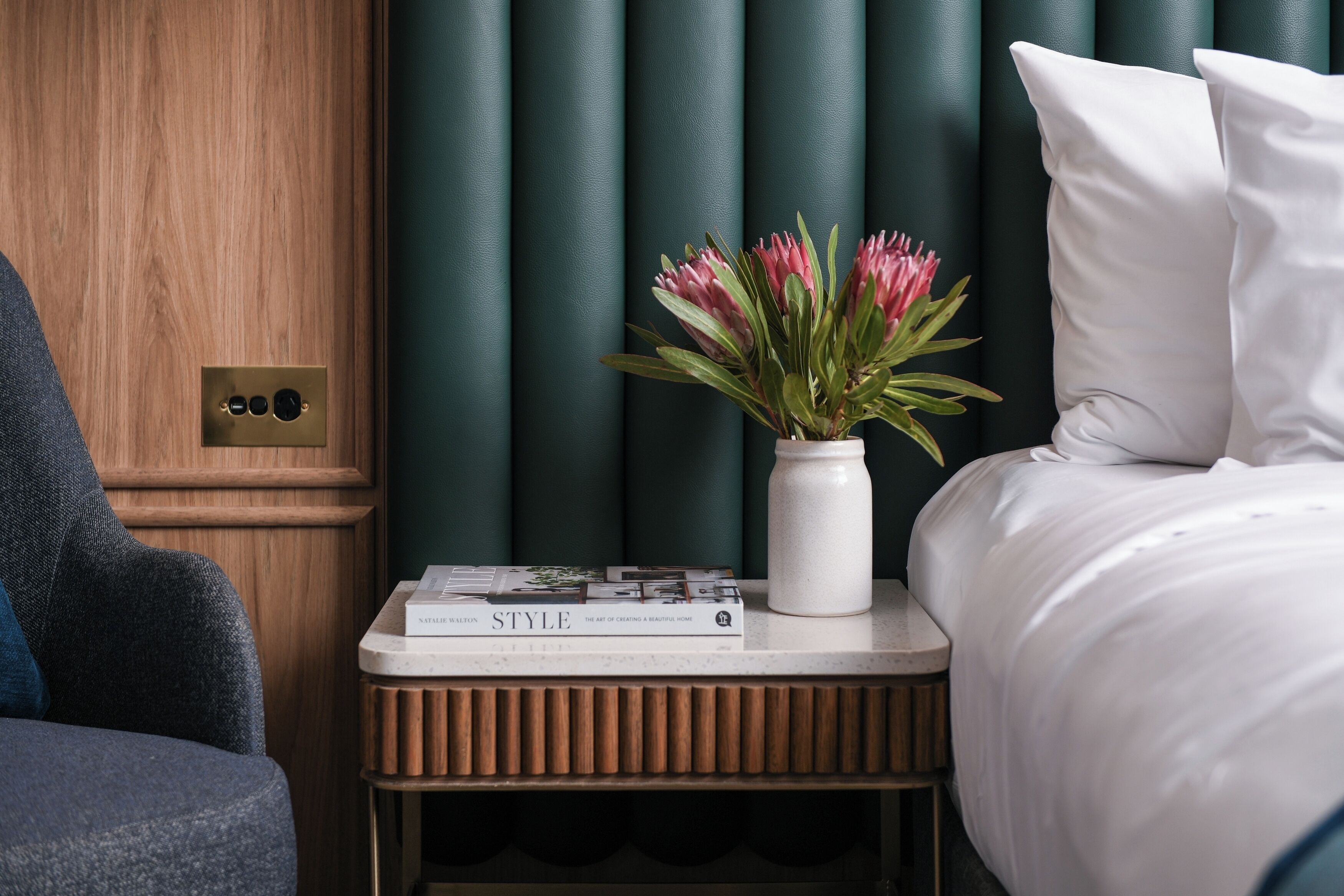 Ein ordentlich eingerichtetes Schlafzimmer mit Holztür, Nachttisch mit Buch und Blumen in einer Vase, sowie einem Bett mit weißer Bettwäsche.