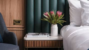 Ein ordentlich eingerichtetes Schlafzimmer mit Holztür, Nachttisch mit Buch und Blumen in einer Vase, sowie einem Bett mit weißer Bettwäsche.