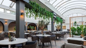 A spacious indoor dining area with multiple tables and chairs, a high ceiling with a skylight, and greenery hanging from the ceiling. - NH Eindhoven Conference Centre Koningshof (Veldhoven)