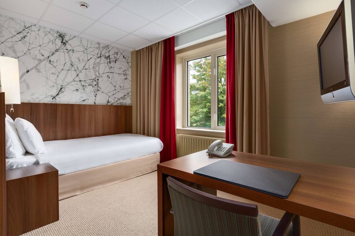 A hotel room with a bed, a desk with a telephone, a chair, a television, and a window with curtains.