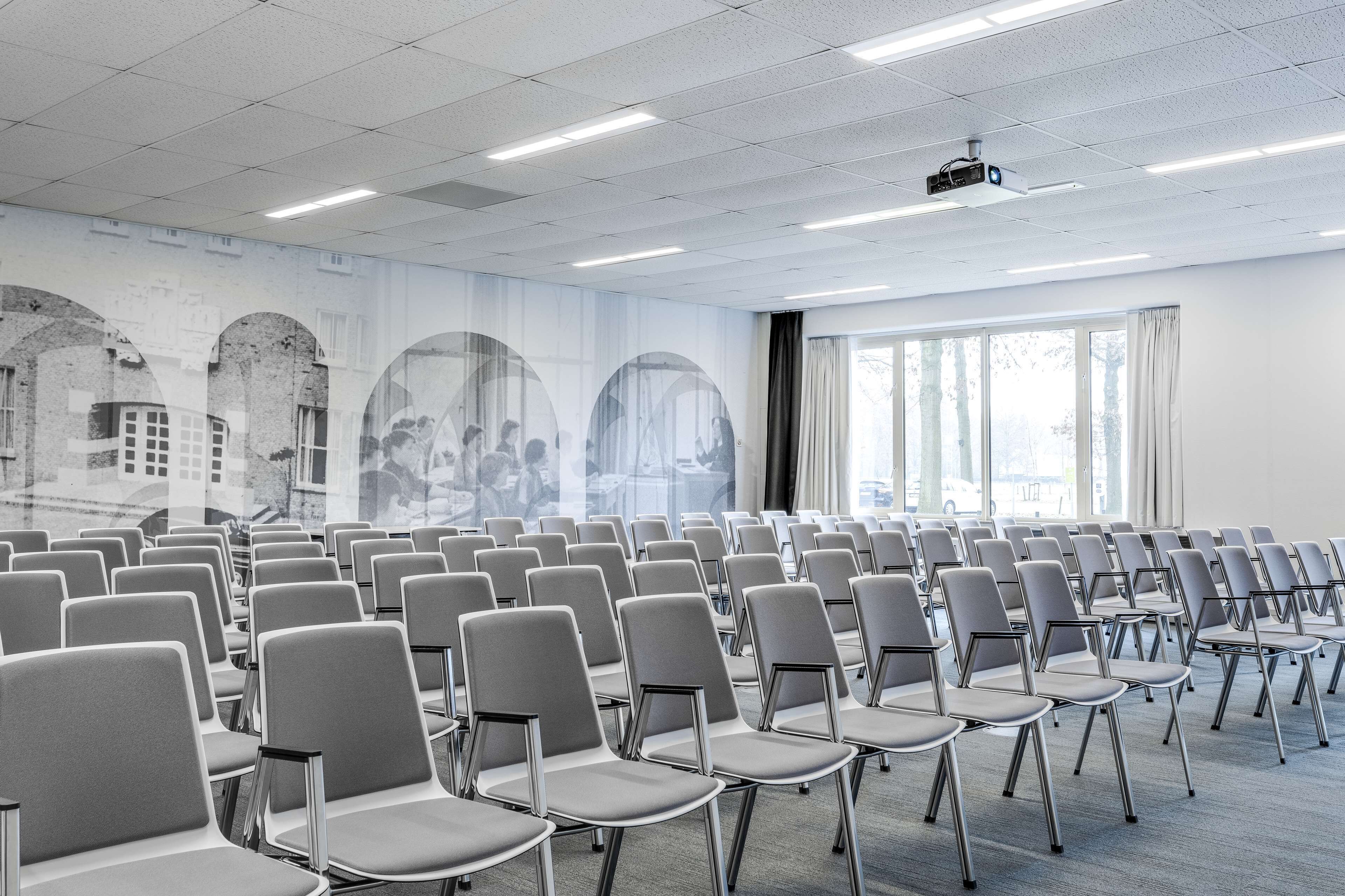 A conference room with rows of grey chairs, a projector on the ceiling, and large windows with curtains.