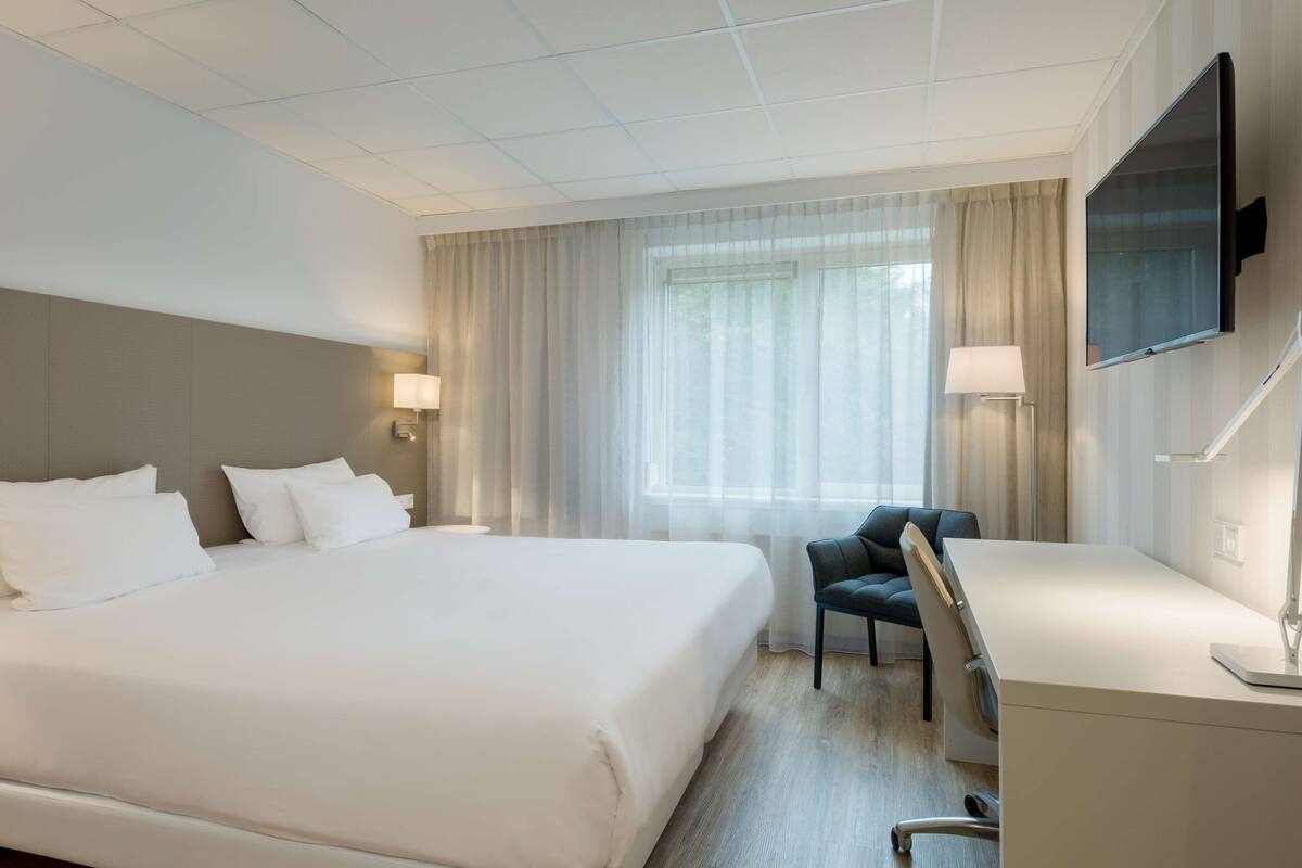 A hotel room with a large bed, a desk, a chair, a TV mounted on the wall, and a window with curtains.