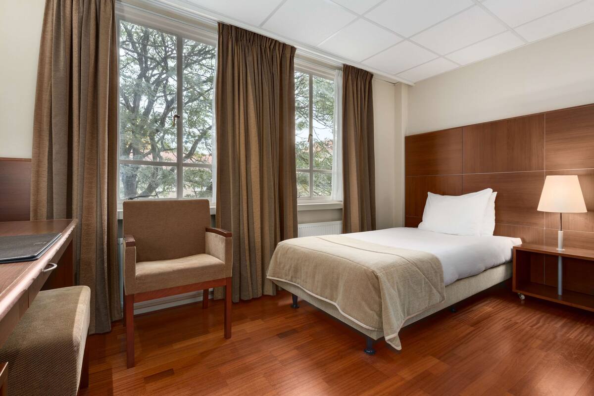 A hotel room with a bed, a desk, a chair, and a nightstand with a lamp.