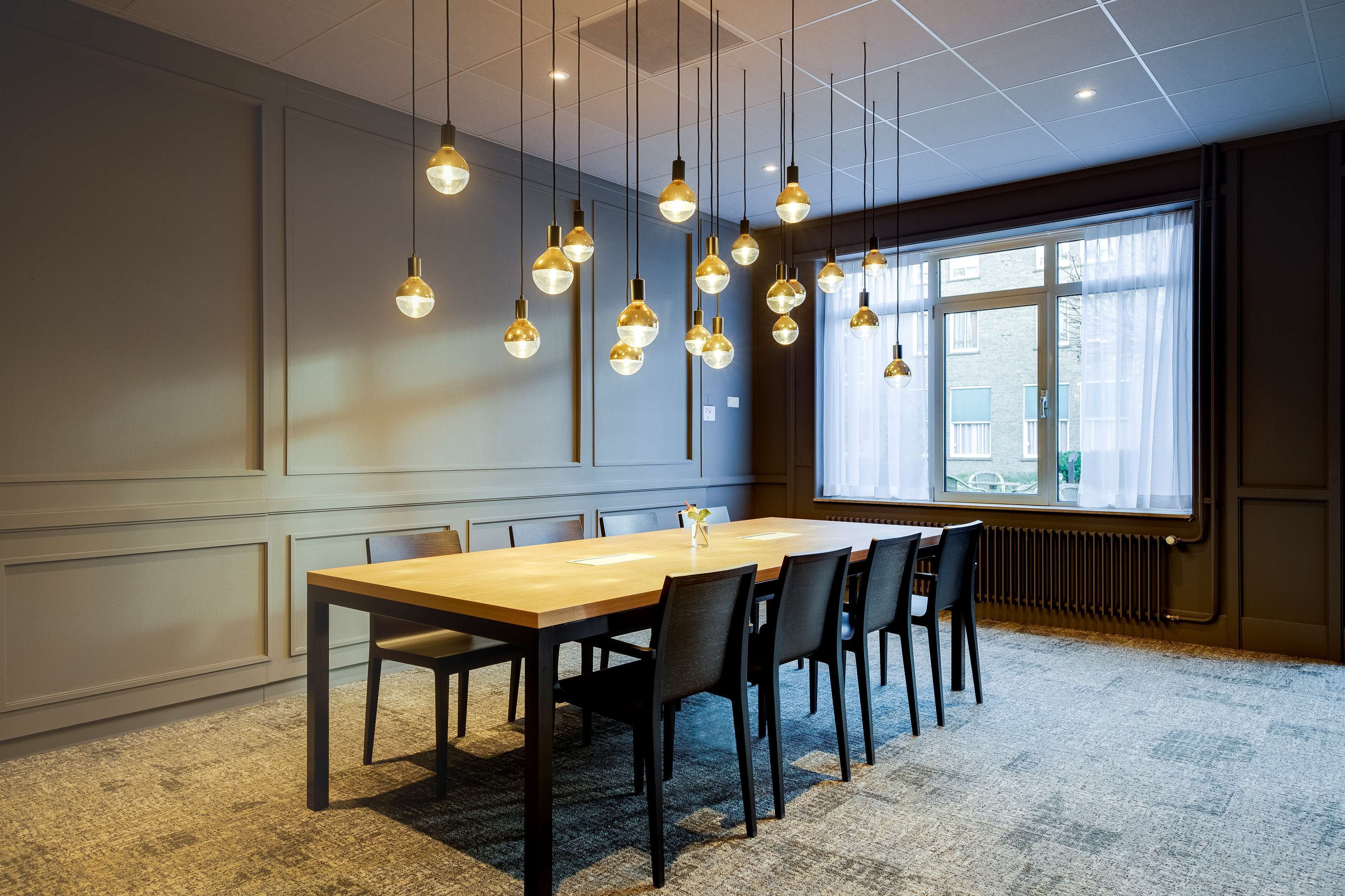 A conference room with a long table, multiple chairs, hanging pendant lights, a large window with curtains, and a radiator.