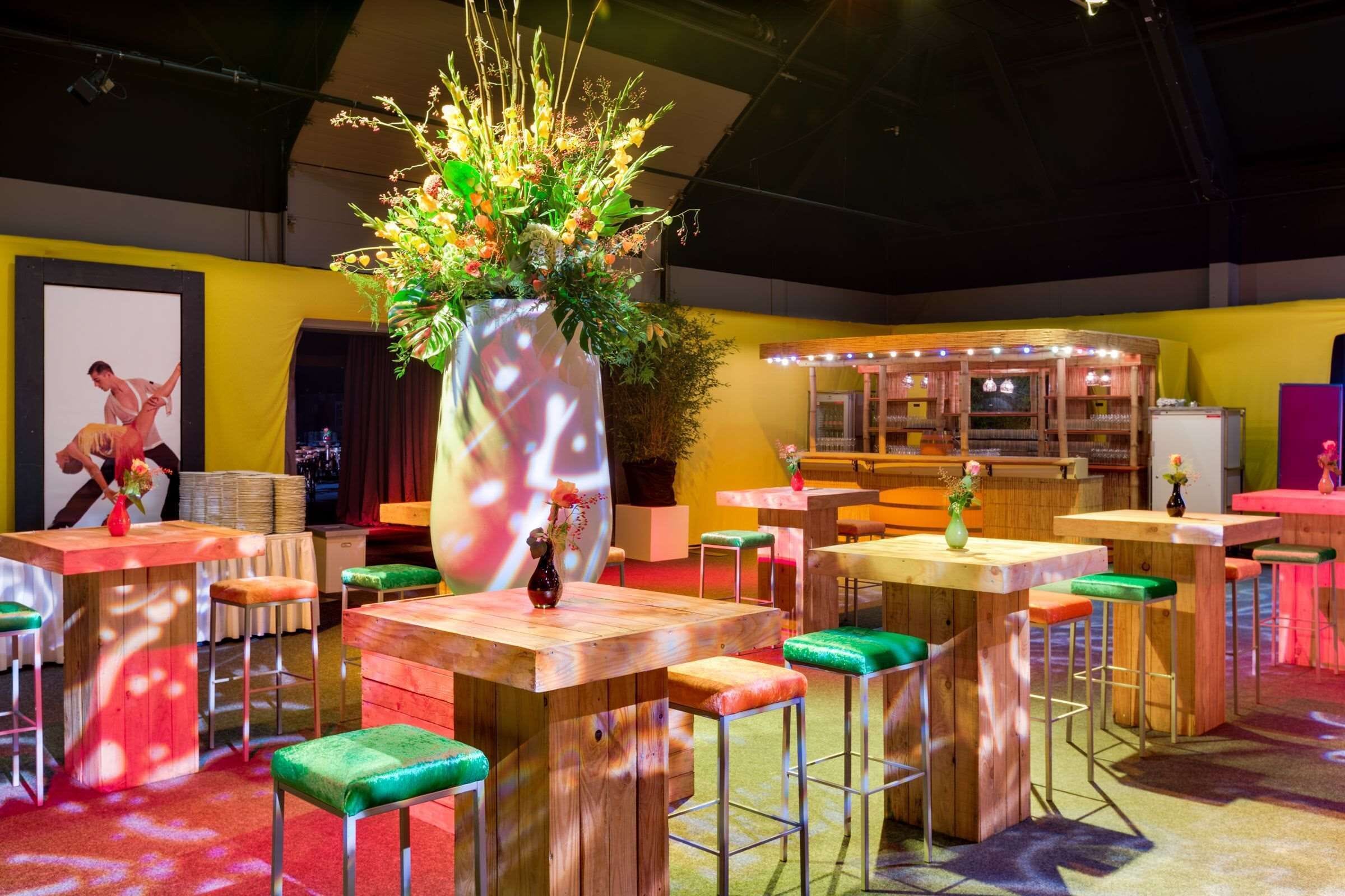 Un espace intérieur accueillant, avec des tables en bois et des tabourets rembourrés de couleur verte, un grand arrangement floral en guise de décoration centrale, et un écran de projection sur lequel est diffusée l’image d’une personne.