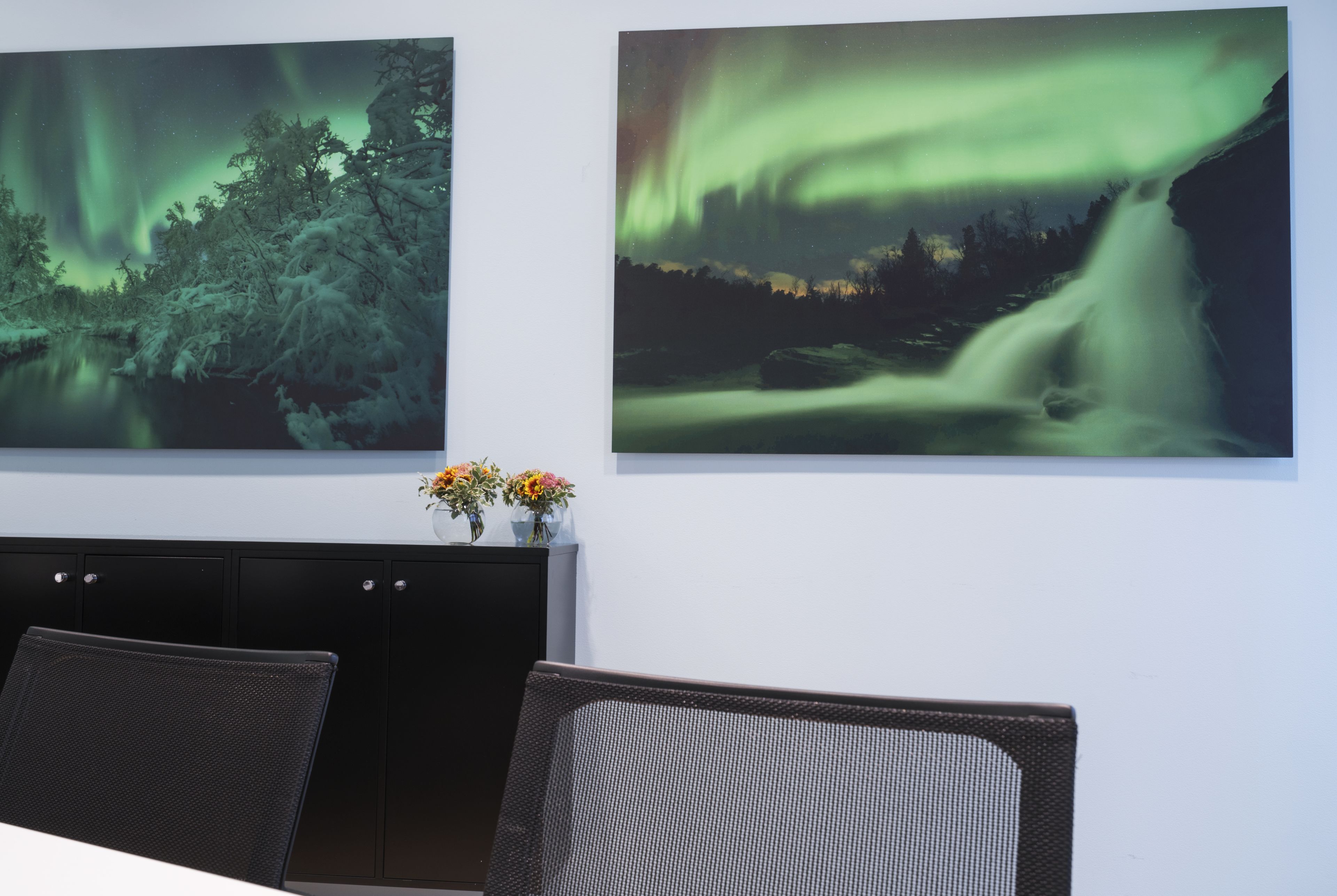 Una habitación con dos ventanas amplias que muestran un bosque nevado y una cascada bajo la aurora boreal. Se observa una cama con cabecera negra, una pequeña mesa con flores y un mueble de color oscuro.
