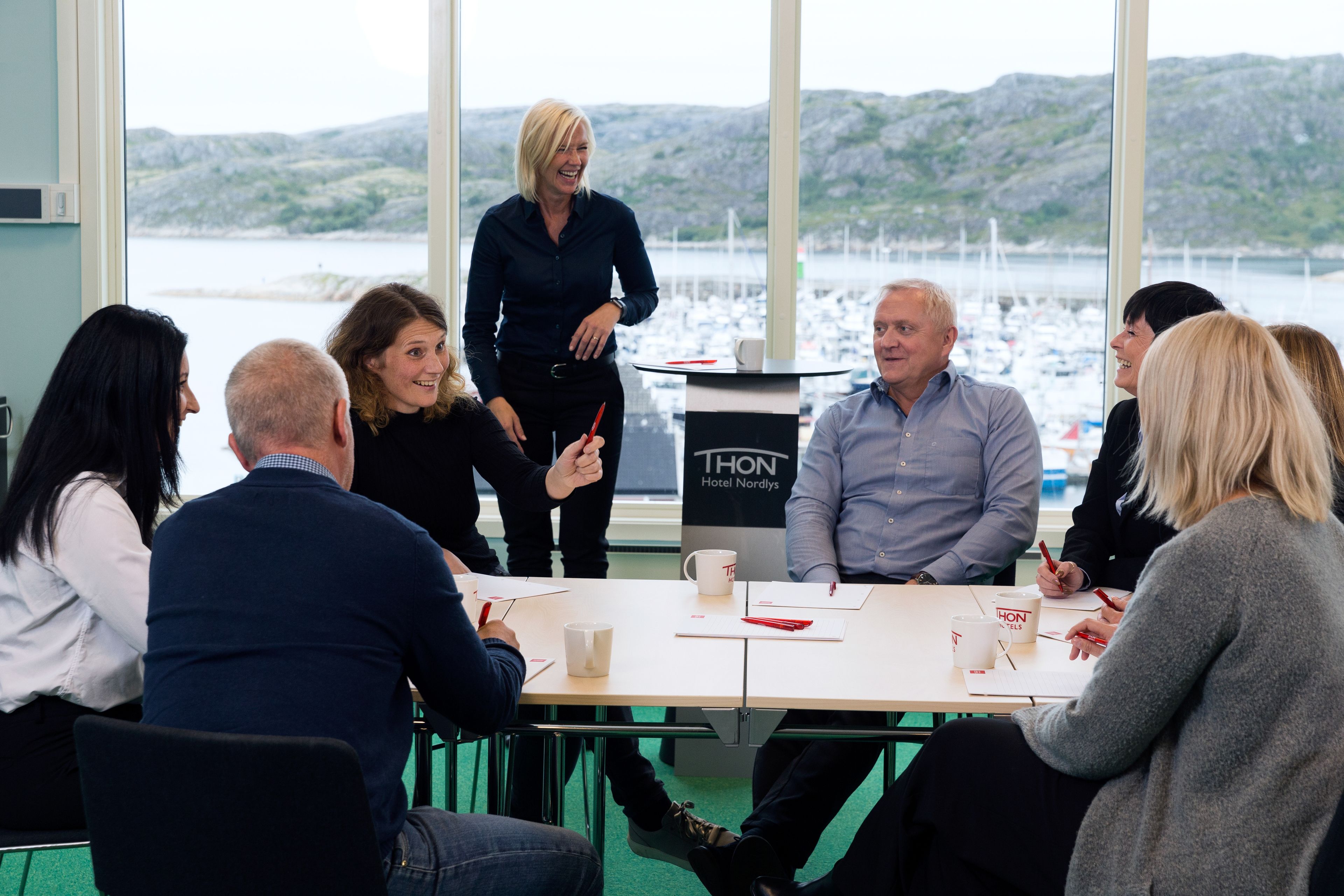 A group of people in a meeting room with a view of a marina and hills in the background.