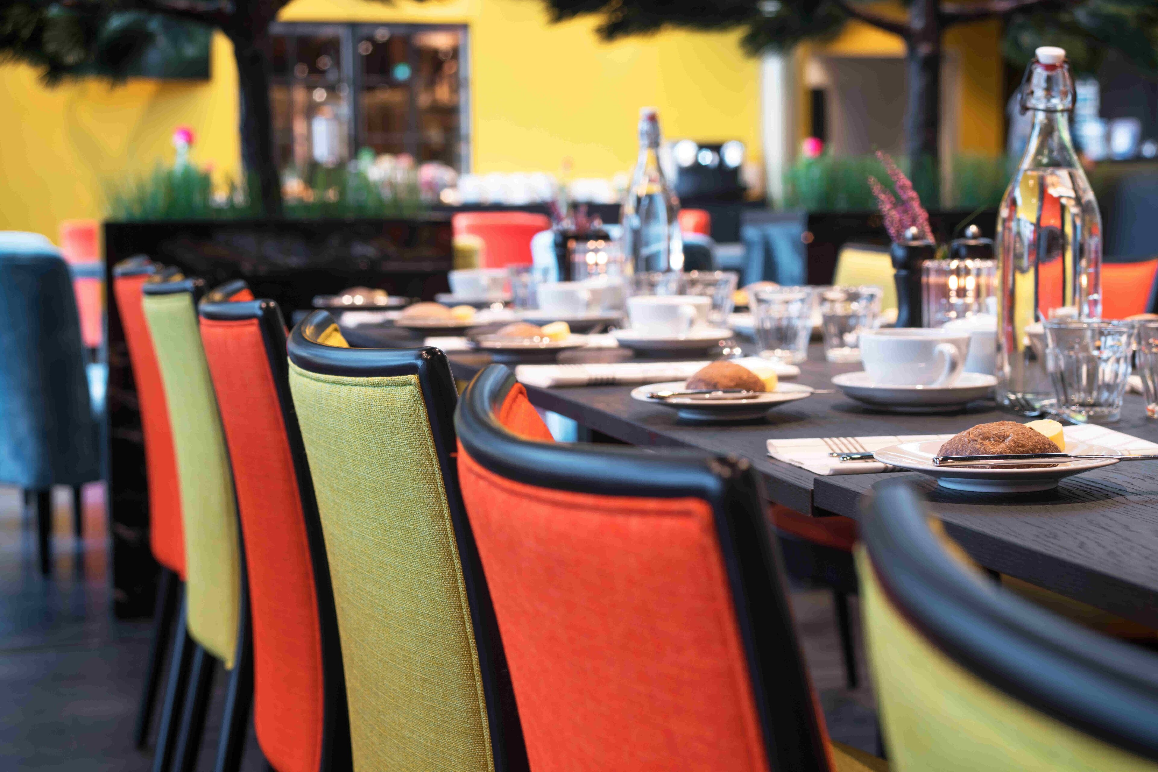 Une table à manger dressée avec des assiettes, des verres et une bouteille, entourée de chaises aux couleurs vives.