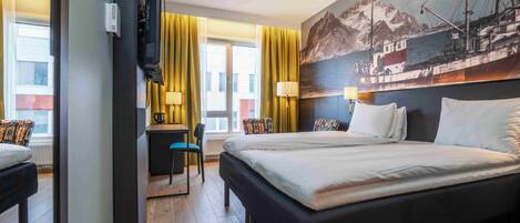 Une chambre d’hôtel avec un grand lit, un bureau avec une chaise, une télévision et un tableau représentant un bateau accroché au mur.