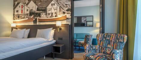 A hotel room with a bed, a nightstand, a chair, a painting of a coastal scene, and a mirror.
