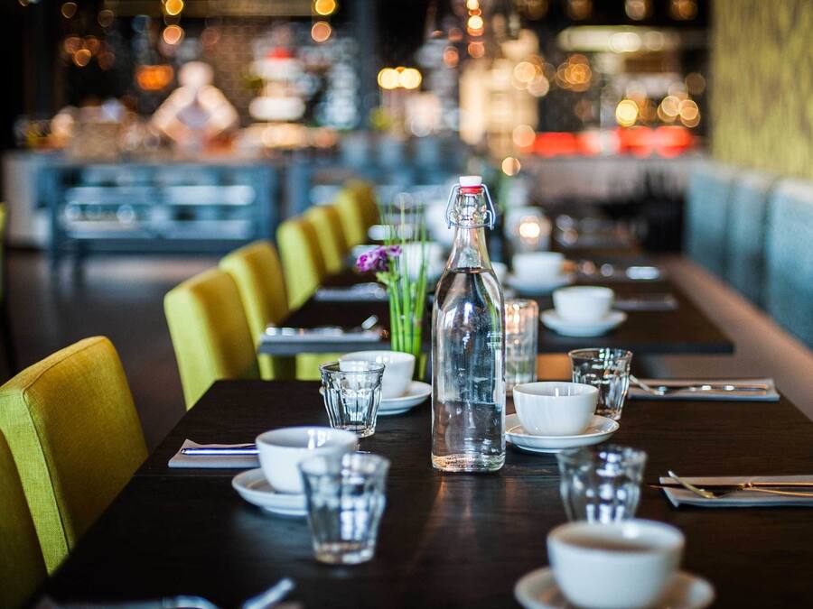 A restaurant table set with a bottle, glasses, and plates, with a blue upholstered bench and yellow chairs in the background.