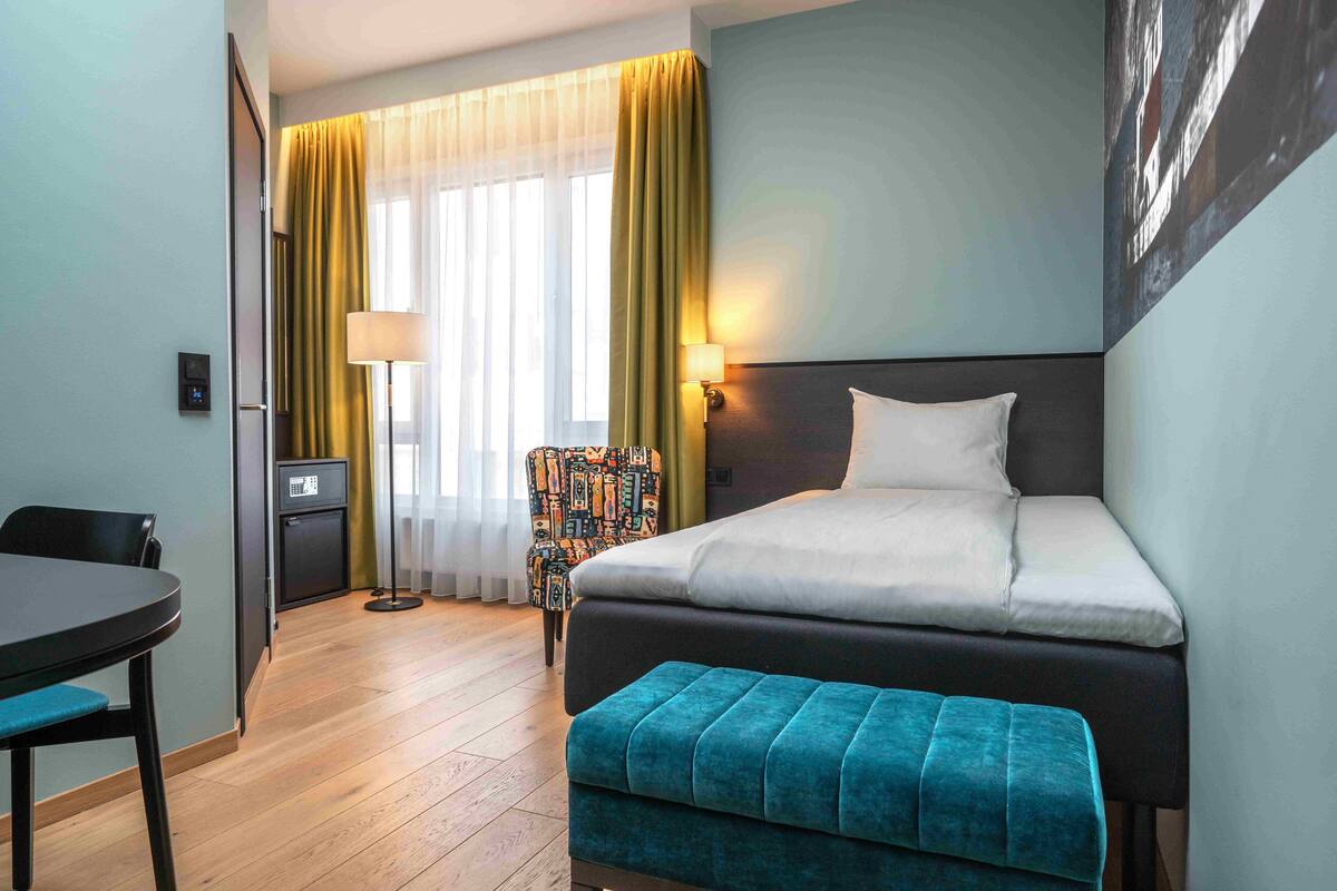 A hotel room with a bed, a desk, a chair, a small kitchenette, and a window with curtains.