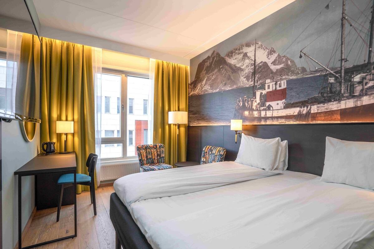 Una habitación de hotel con una cama, dos sillas, un escritorio y un ventanal con cortinas amarillas.