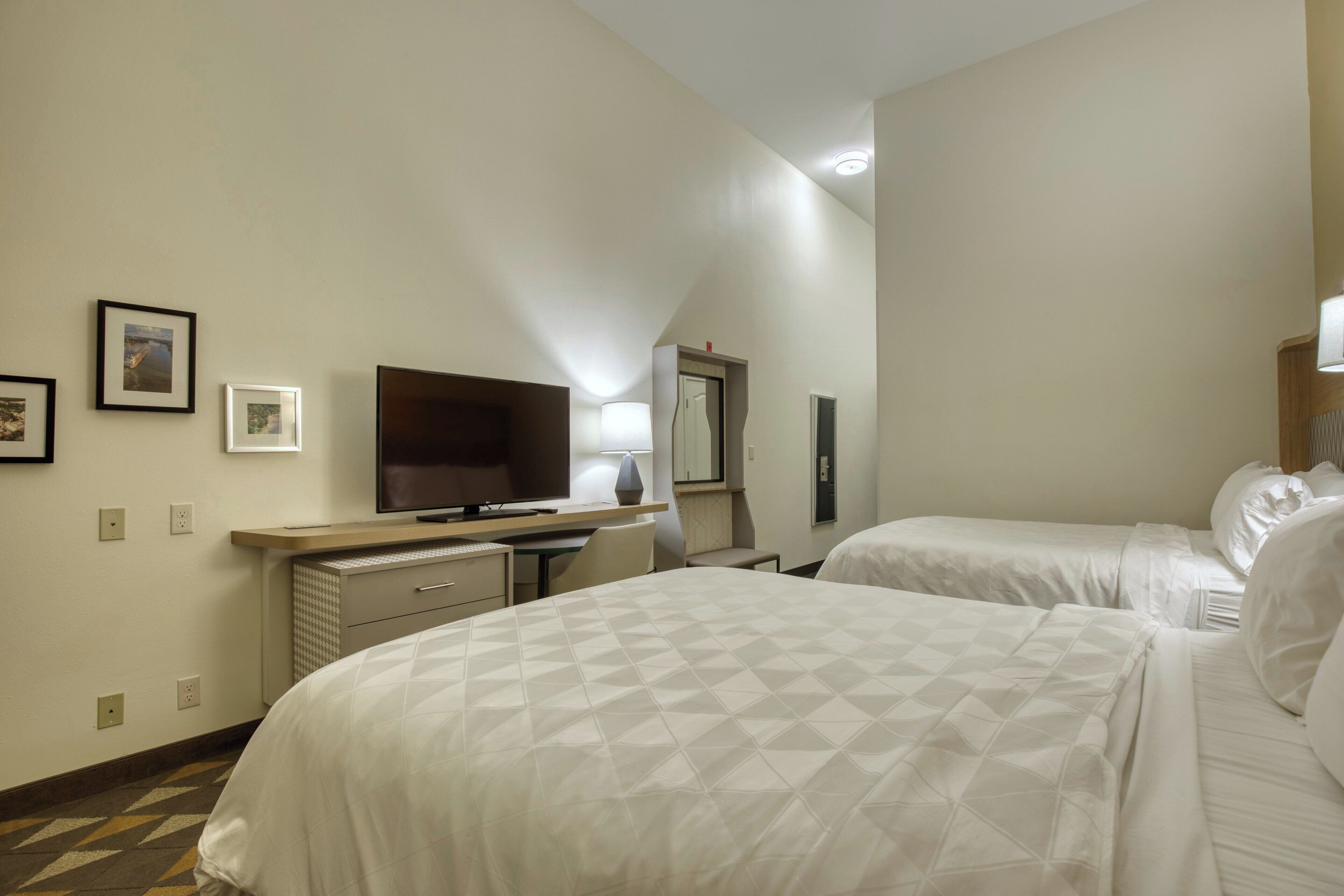 A hotel room with two beds, a desk with a flat-screen TV, a dresser, and a mirror.