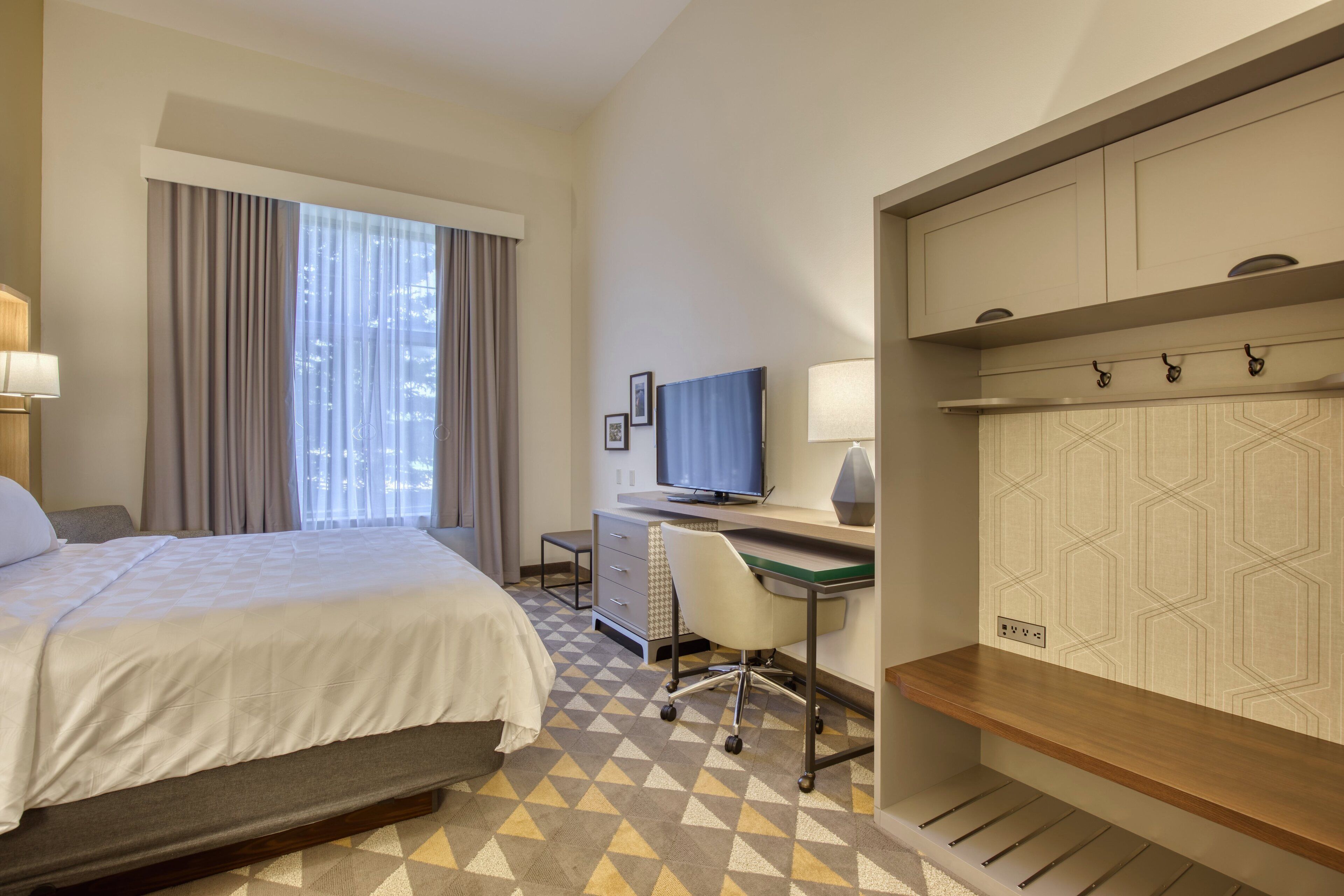 Habitación de hotel con cama, escritorio, silla, televisor y vista a la ciudad desde la ventana.