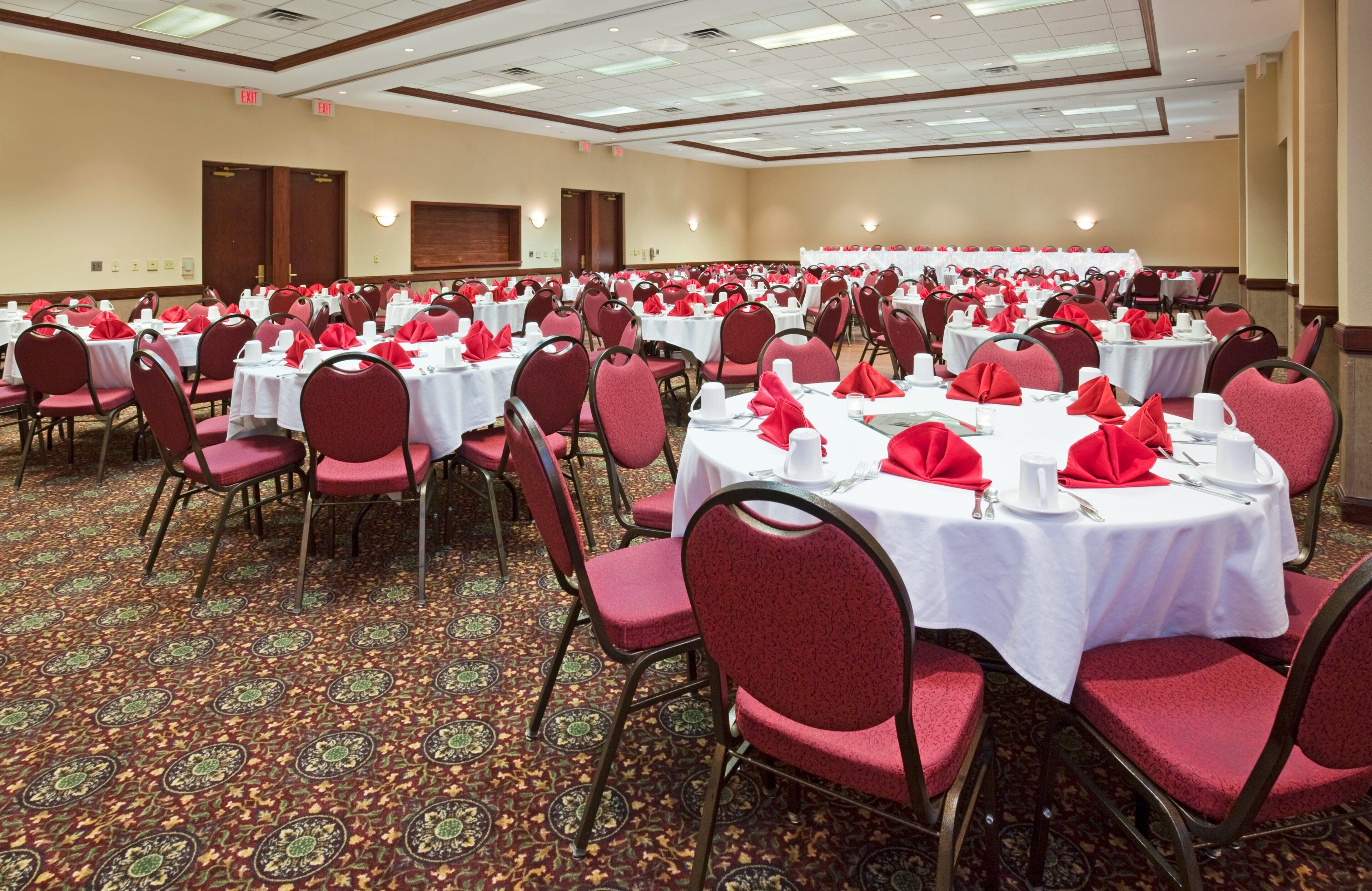 Un salón de banquetes con mesas redondas, sillas rojas y manteles blancos.