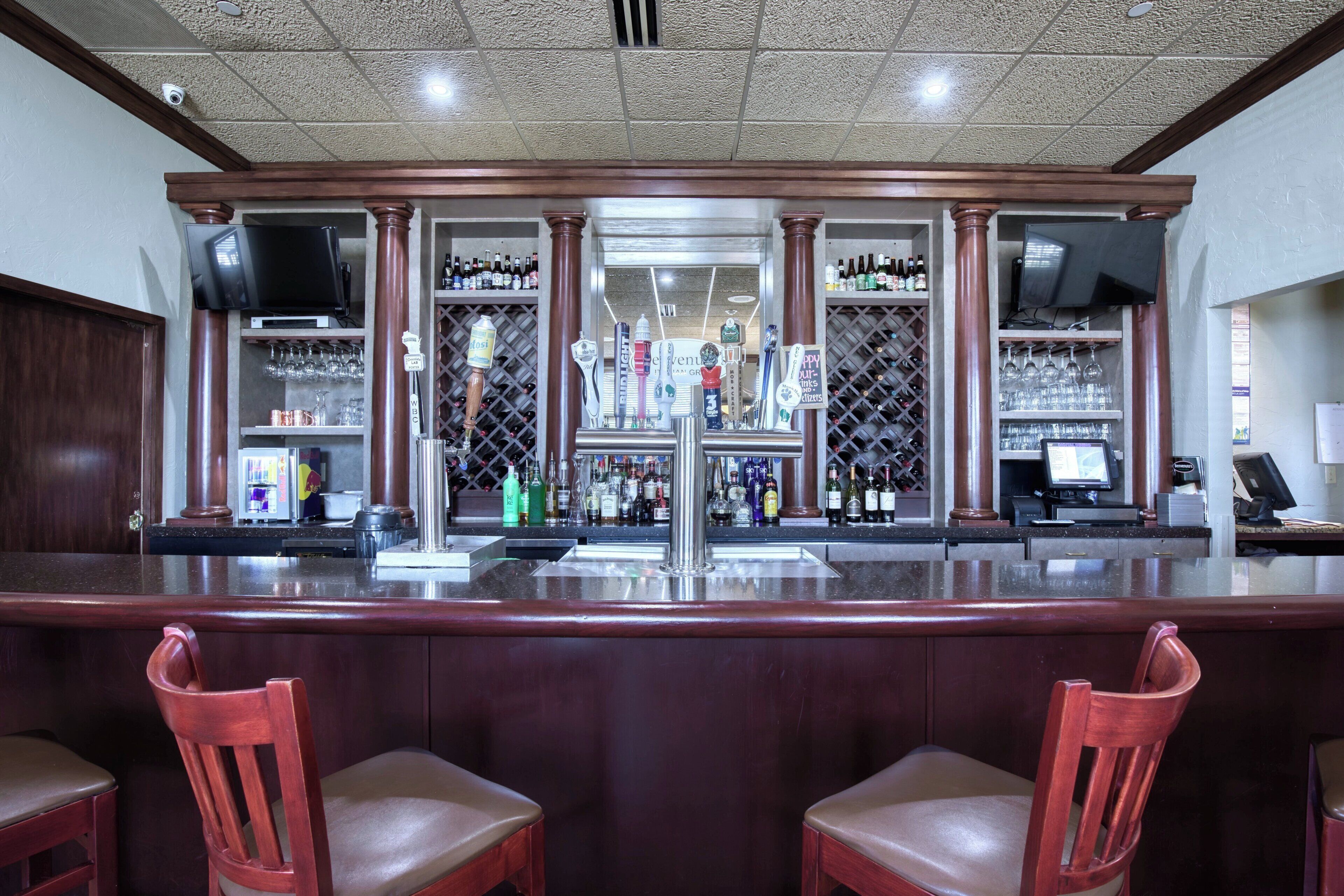 A bar area with a counter, multiple taps, shelves with bottles, and two wooden chairs.