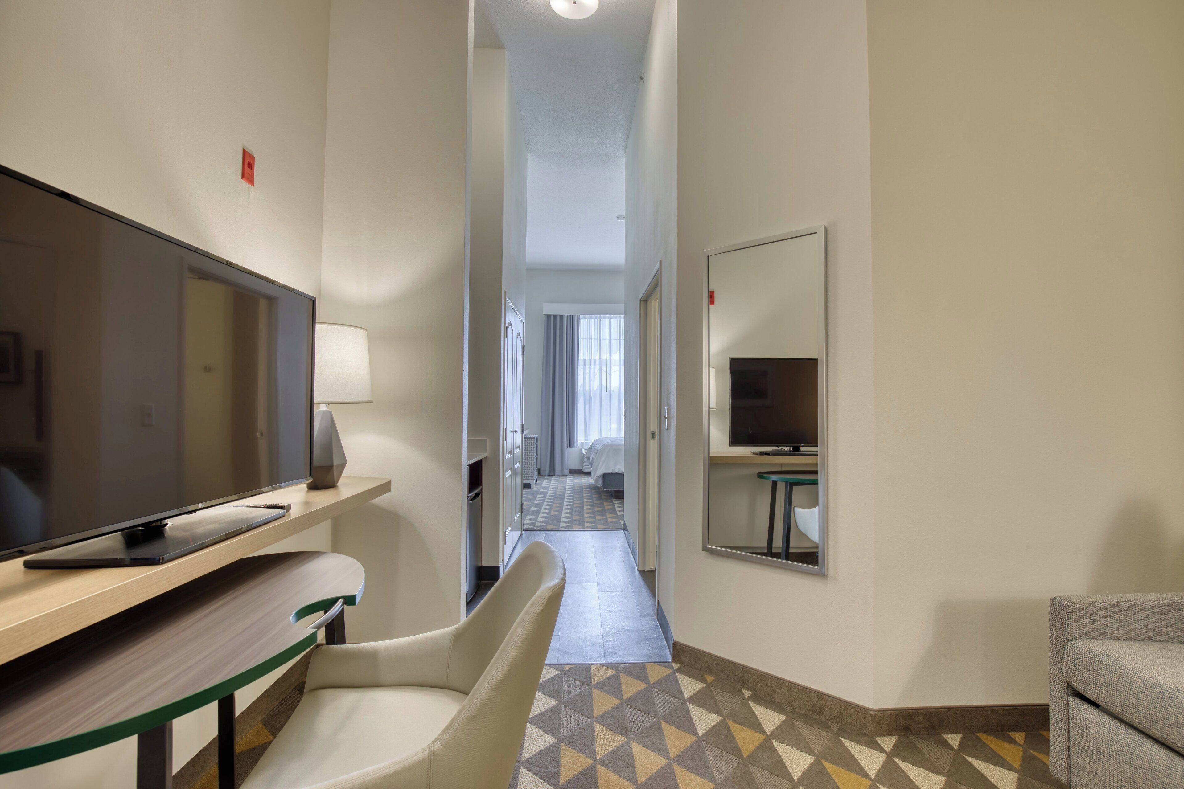 A hotel room with a flat-screen TV, a desk, a chair, a mirror, and a patterned floor.