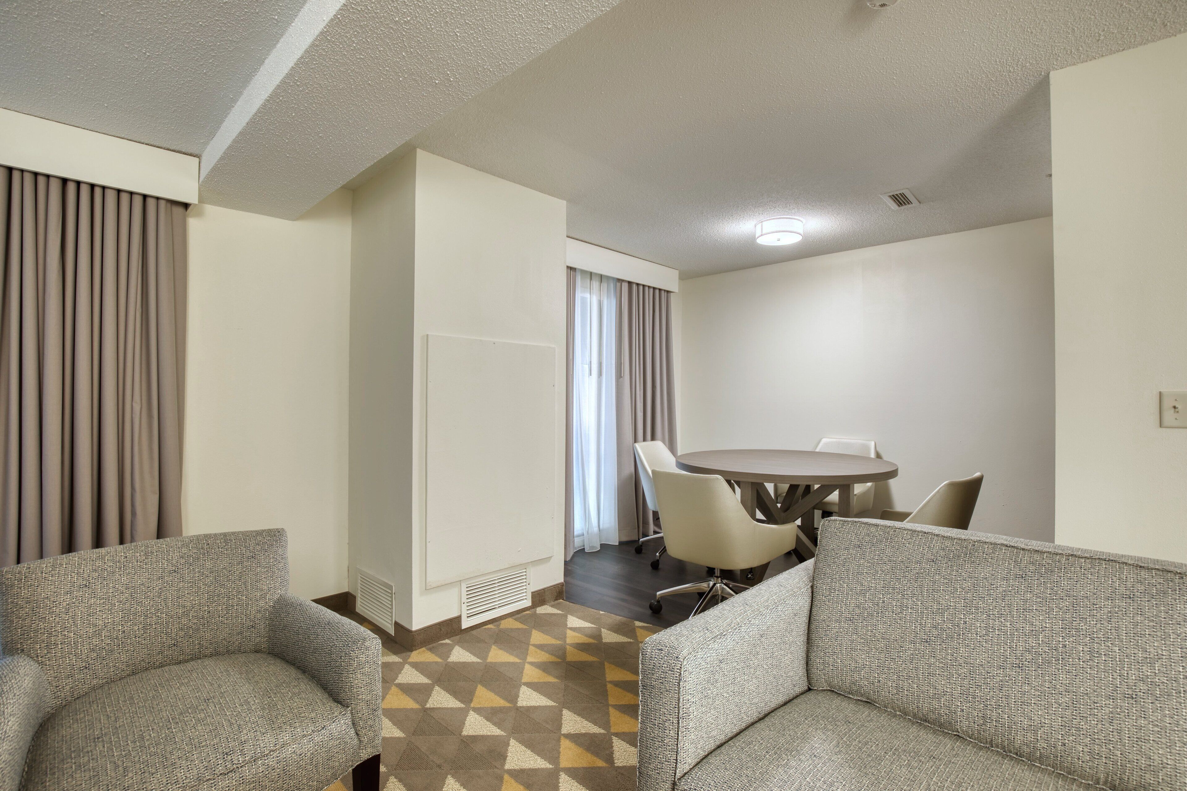 A hotel room with a sofa, a dining table with chairs, a window with curtains, and a patterned floor.