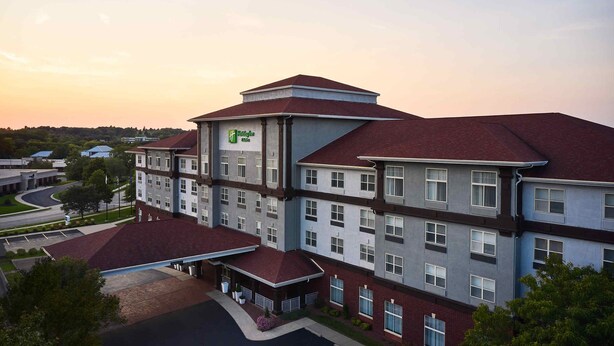 A multi-story hotel with a prominent green logo, a parking lot, and a landscaped area with trees.