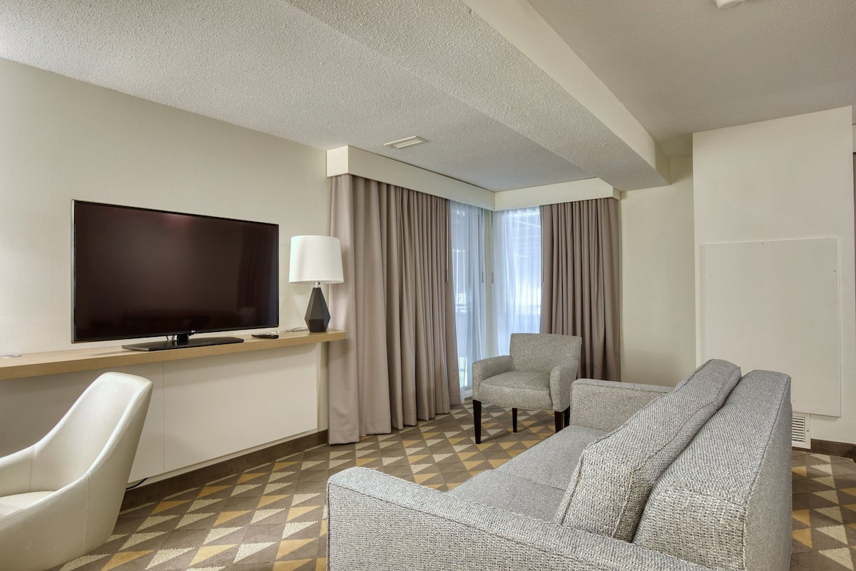 A hotel room with a large flat-screen TV, a sofa, a chair, and a desk with a lamp.