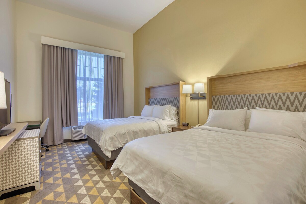 A hotel room with two beds, a desk, a chair, a window with curtains, and a lamp.
